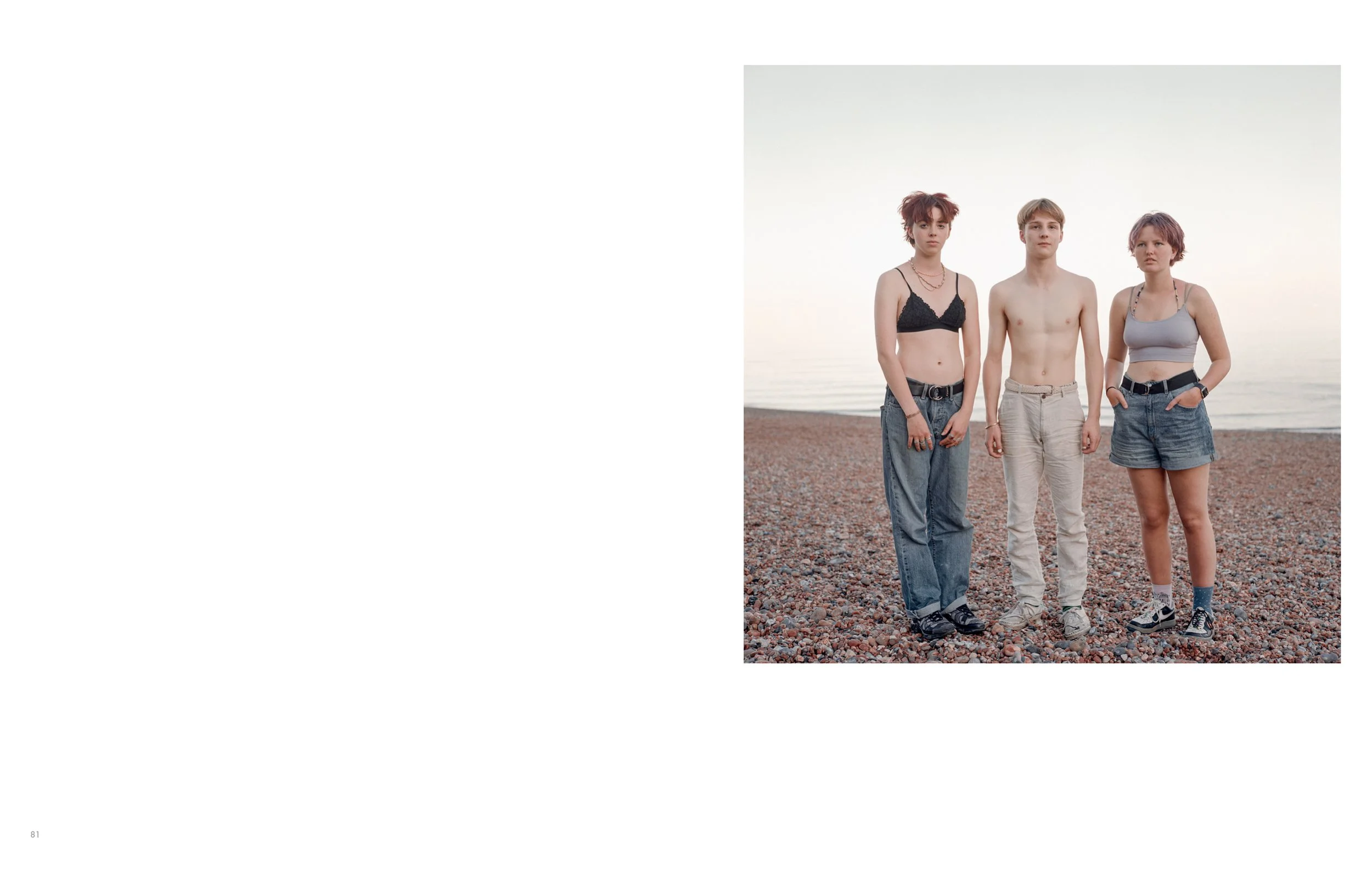 Three young adults standing on a pebbled beach during sunset, wearing casual clothing, with a calm body of water in the background.