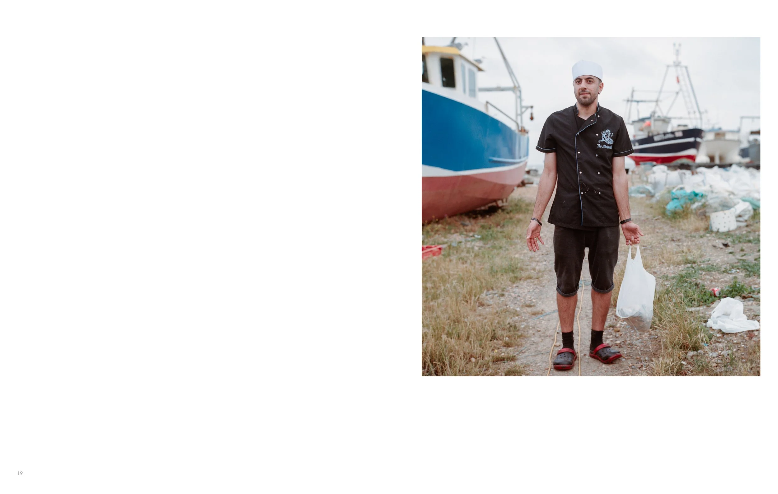 A man standing outdoors near boats at a boatyard, holding a plastic bag, wearing black clothing, a white cap, and Crocs sandals.