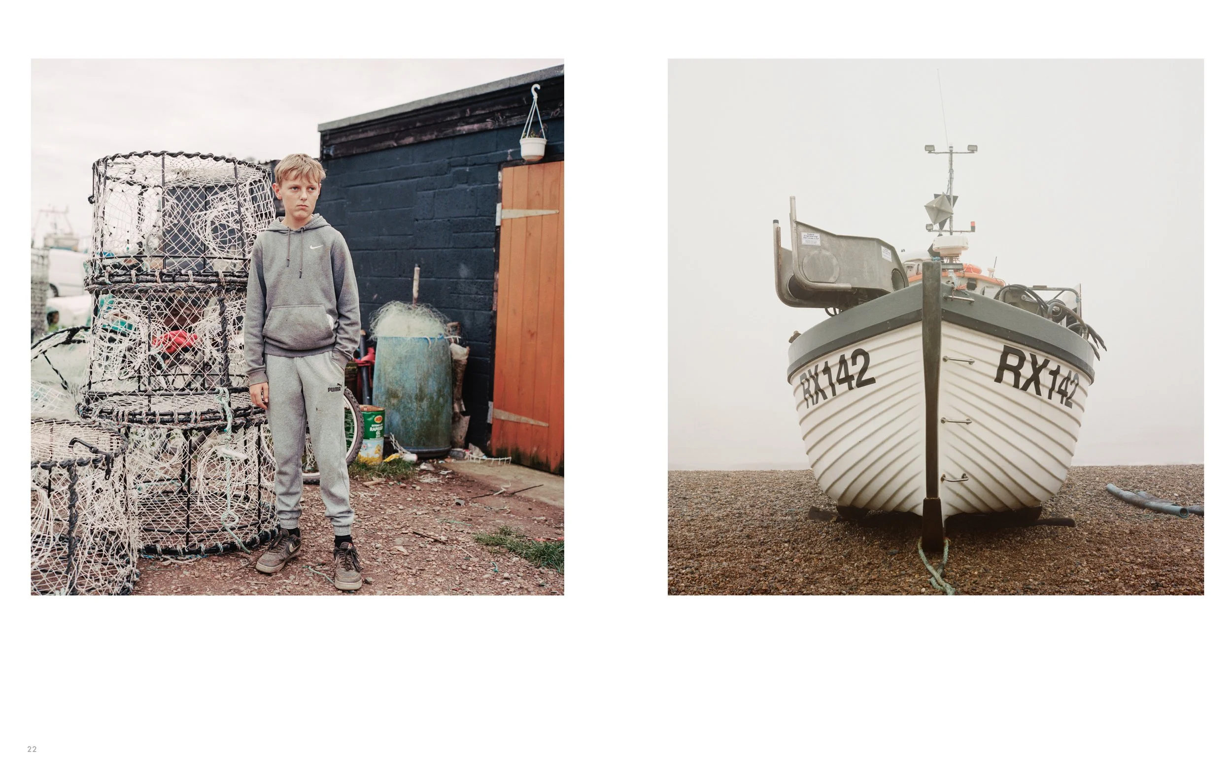 A young boy stands next to a large fishing trap made of wire, wearing a gray Nike hoodie and matching sweatpants, with a dark-colored building and outdoor items behind him. A boat with identification number RX142 is docked on a pebbled shore under an
