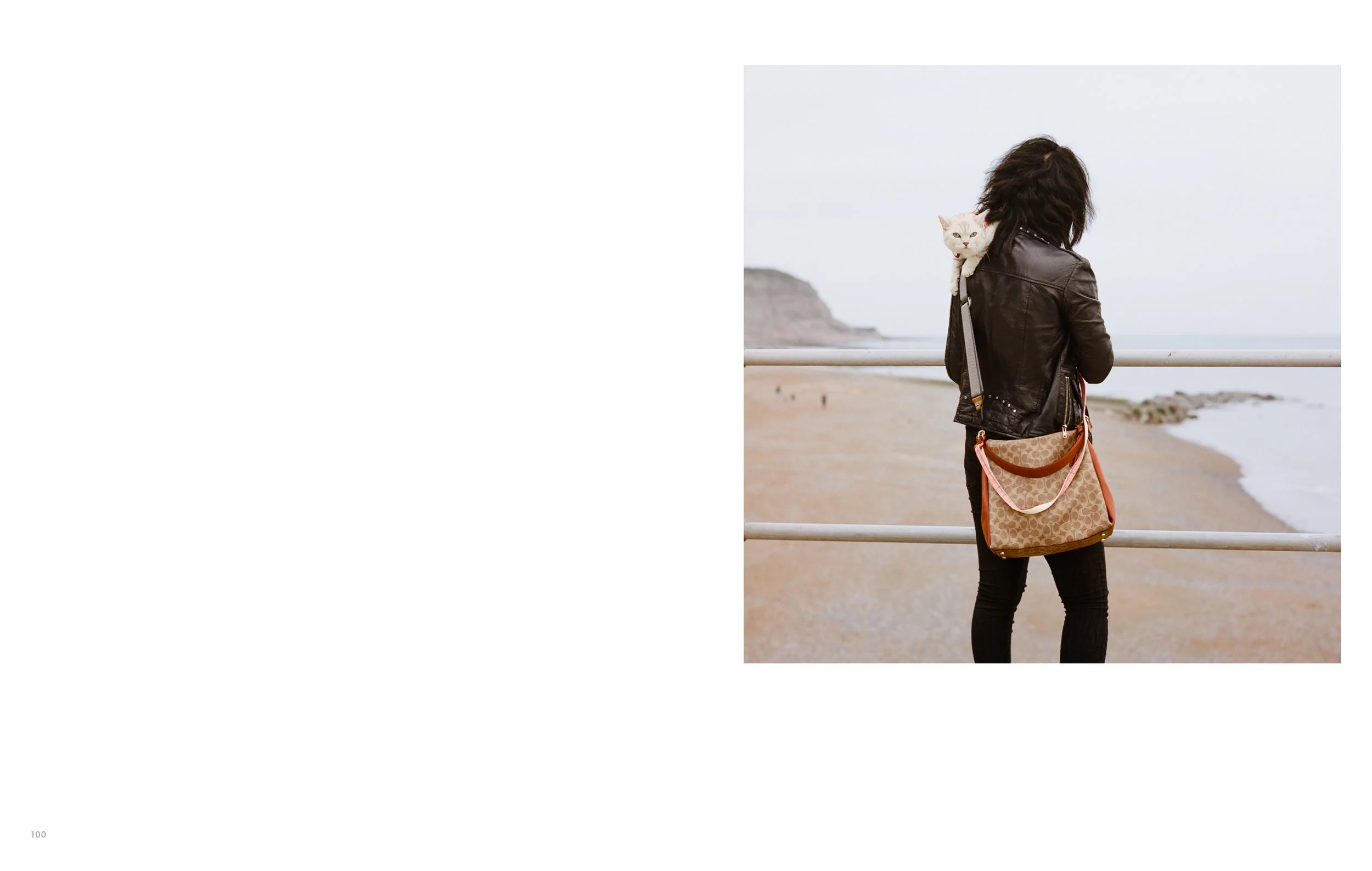 Person with dark curly hair wearing a black leather jacket and carrying a beige bag, standing near a railing and looking out across the beach and ocean.