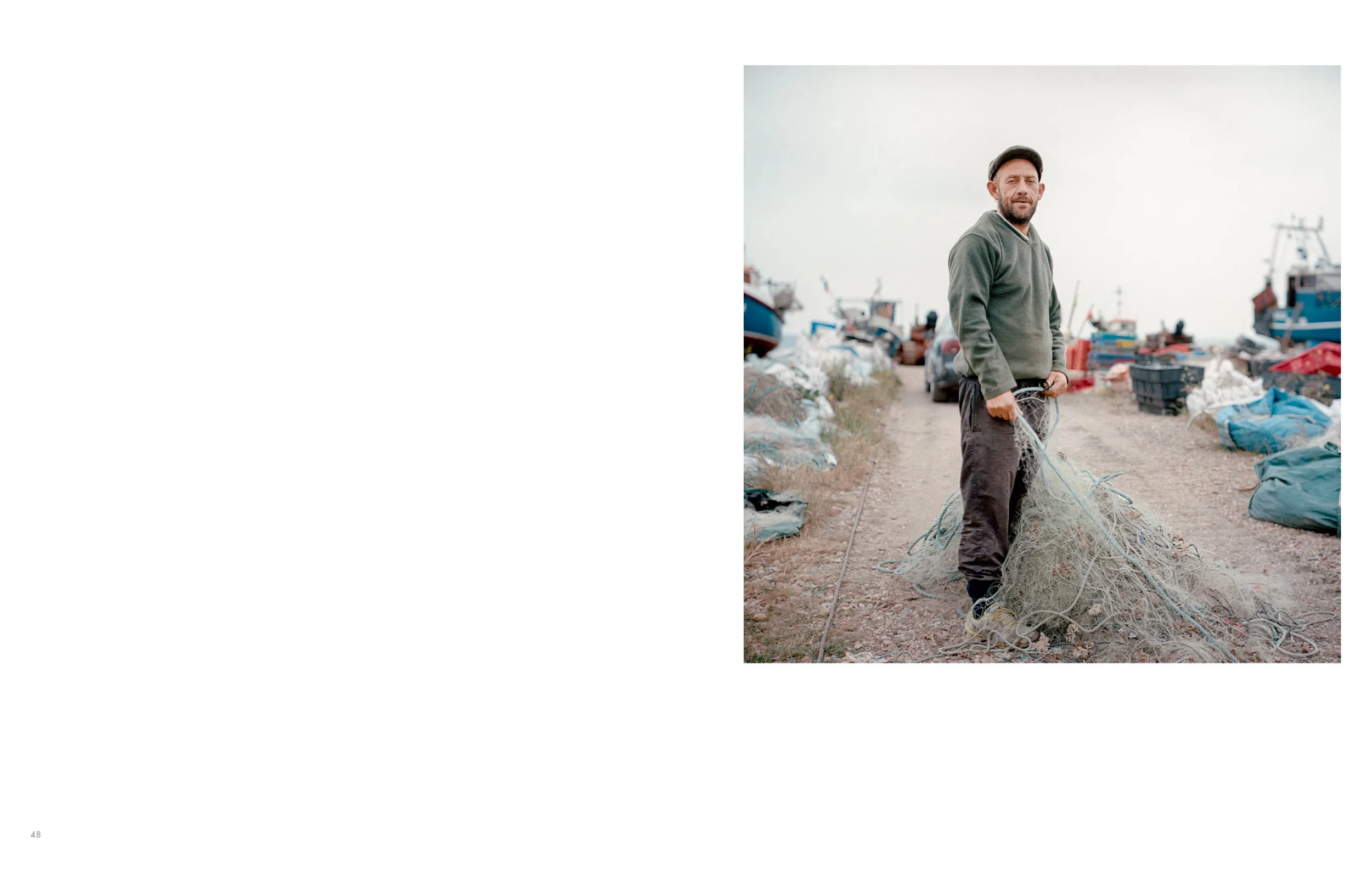 A man standing on a dirt path surrounded by fishing boats and equipment at a harbor, holding tangled fishing net.
