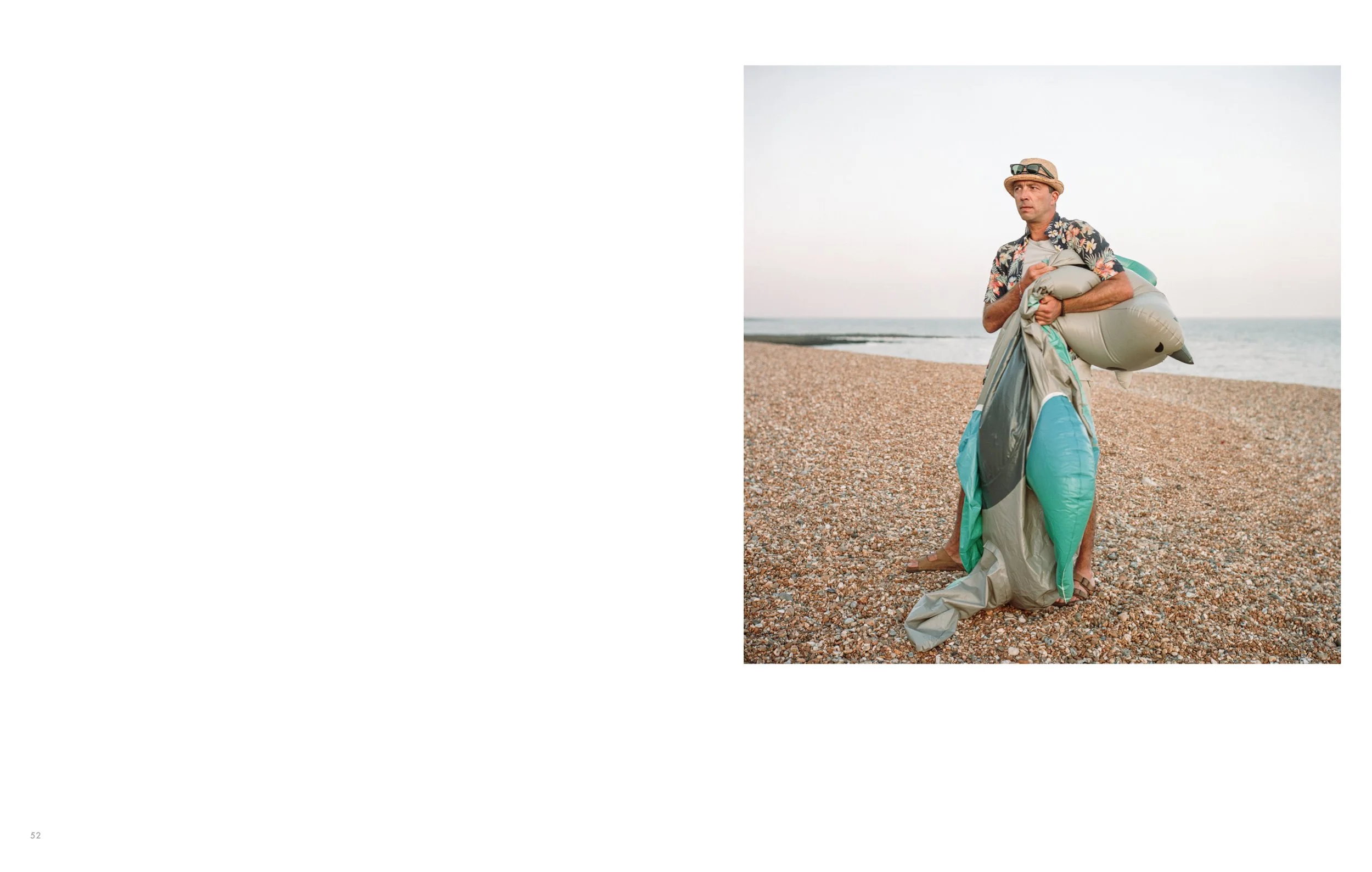 A man on a pebble beach holding an inflatable paddleboard, wearing a hat, sunglasses, a floral shirt, and shorts.