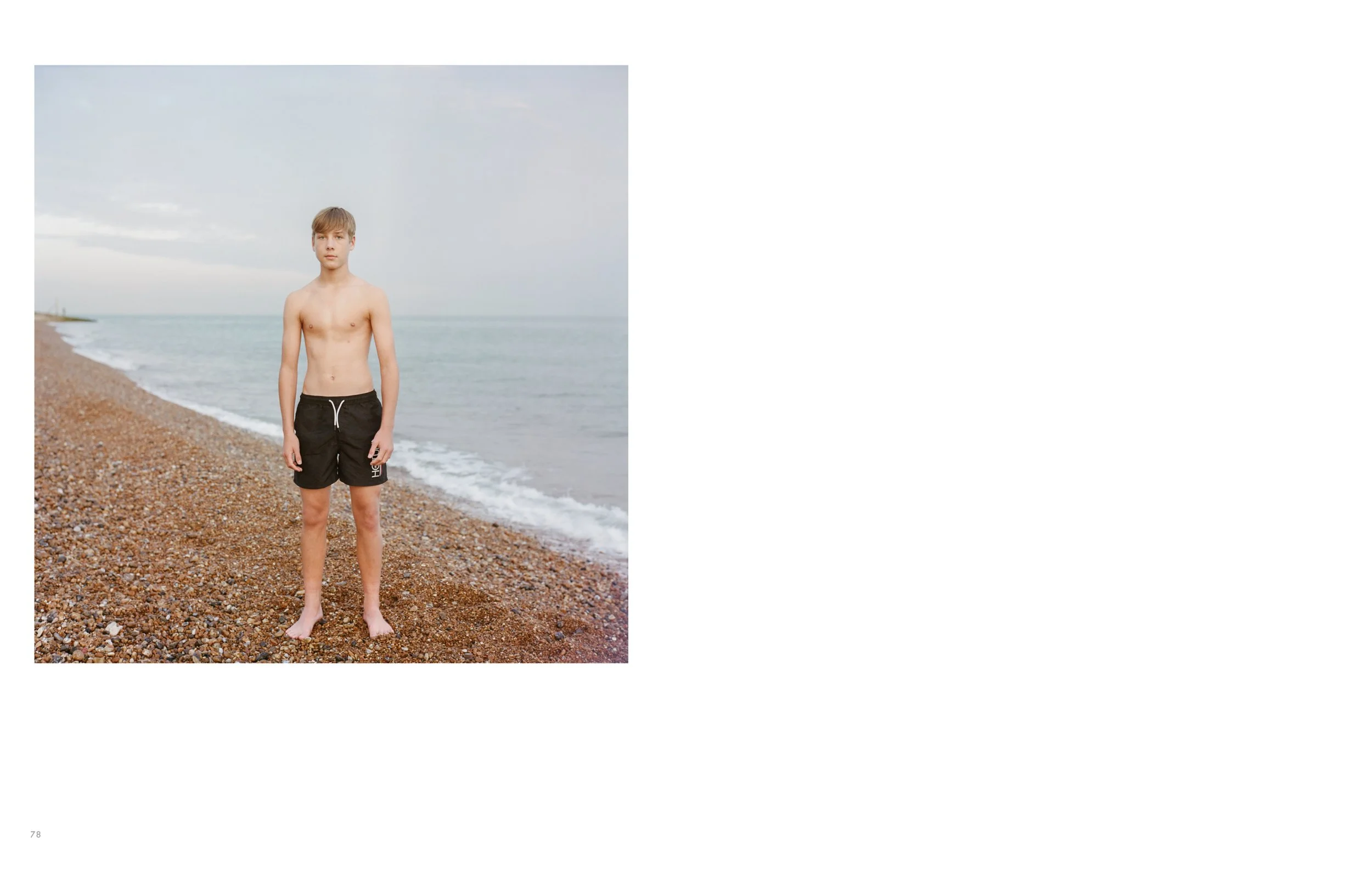 A teenage boy with short blond hair standing barefoot on a pebbled beach near the water, wearing black shorts with a white drawstring, looking at the camera with a neutral expression, overcast sky in the background.