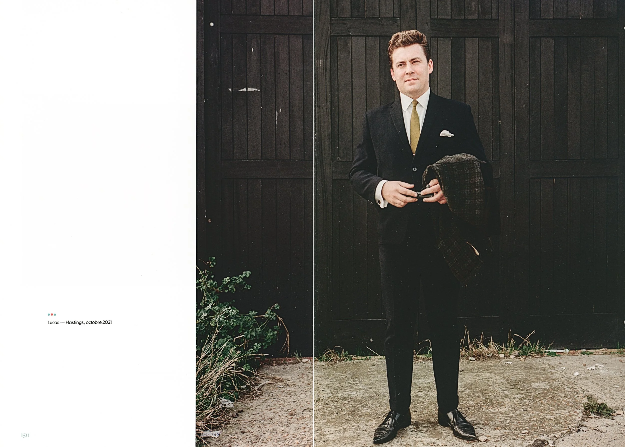 Man in a formal suit standing by a black wooden wall, holding a jacket, with greenery at his feet.