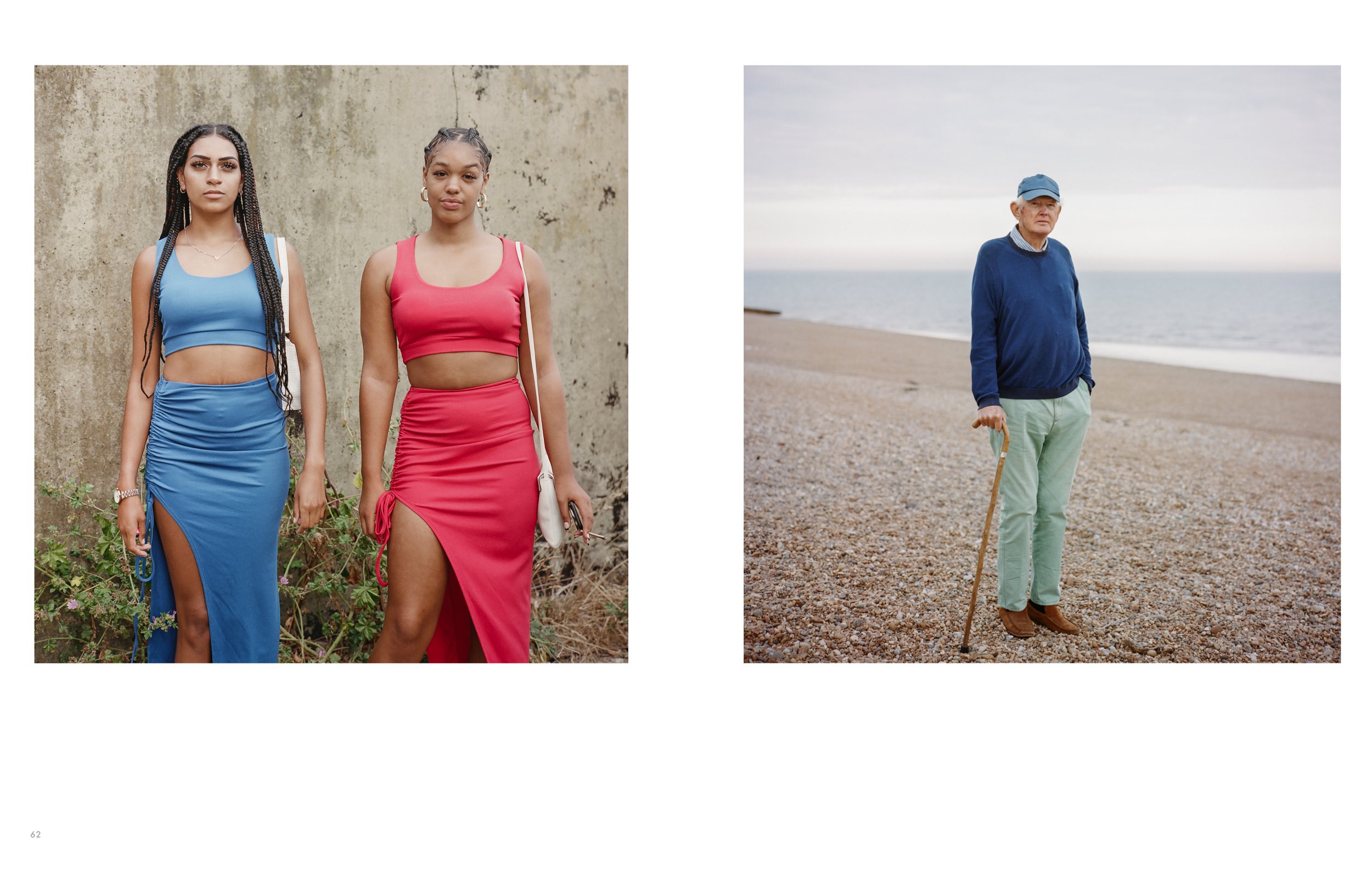 Photo of two young women standing outdoors against a concrete wall on the left, and an older man with a walking stick standing on a pebble beach near the water on the right.