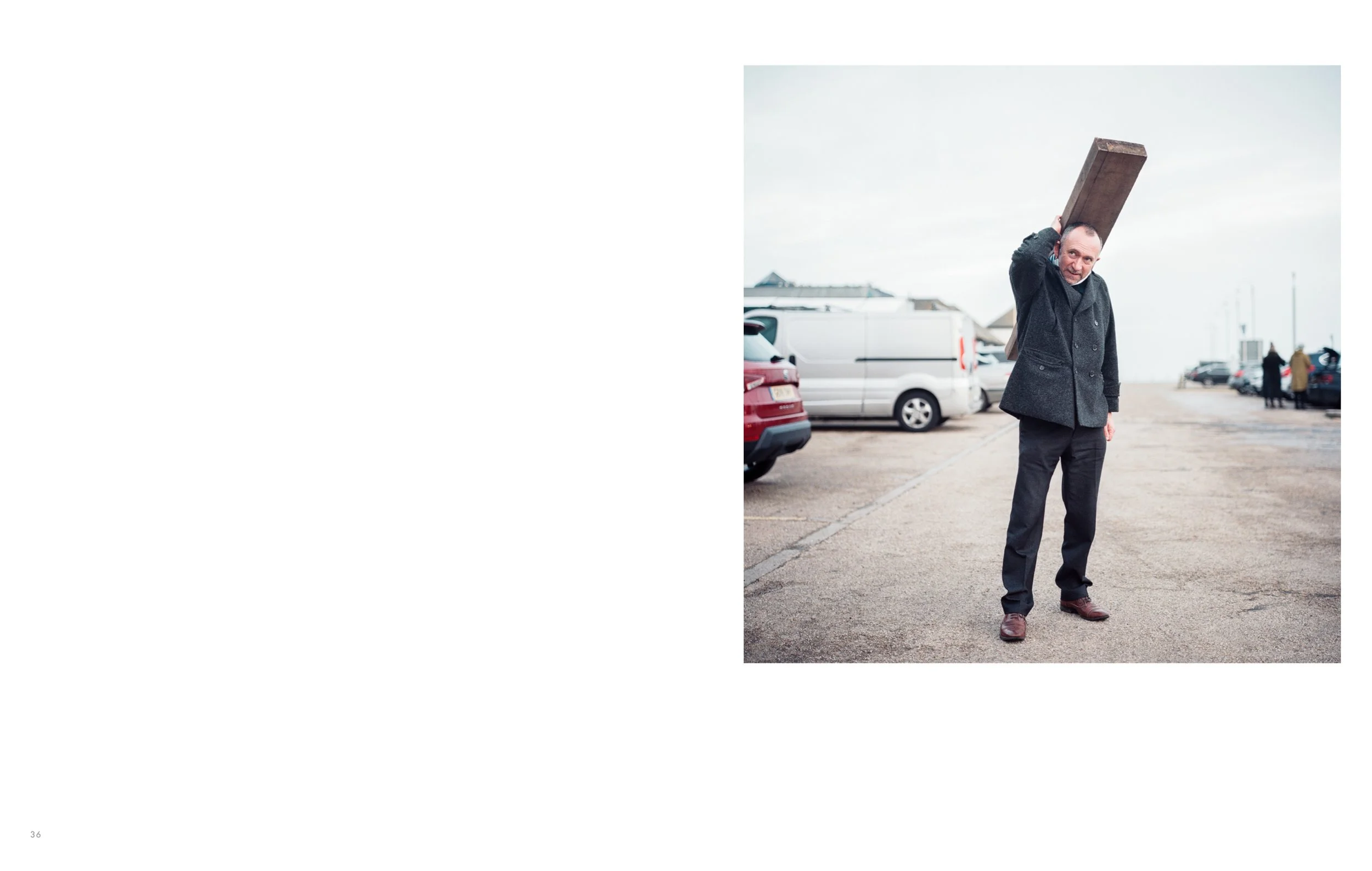 Man in dark coat lifting a large wooden plank onto his shoulder in a parking lot, with cars and people visible in the background.