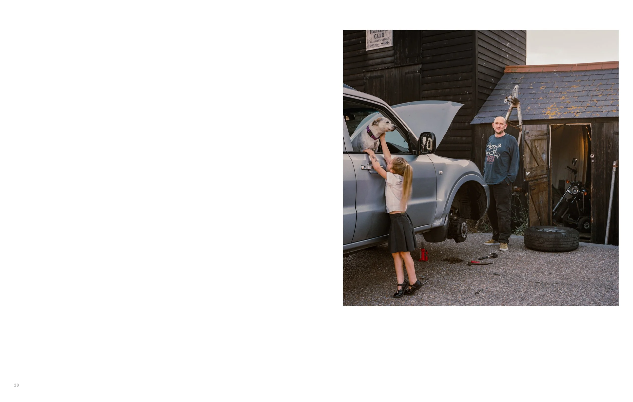 A man and two young girls working on a car in a driveway next to a wooden shed. The man is standing and smiling while a girl points at the car engine. Another girl is reaching into the engine bay of the car. The area has a tire and tools on the groun