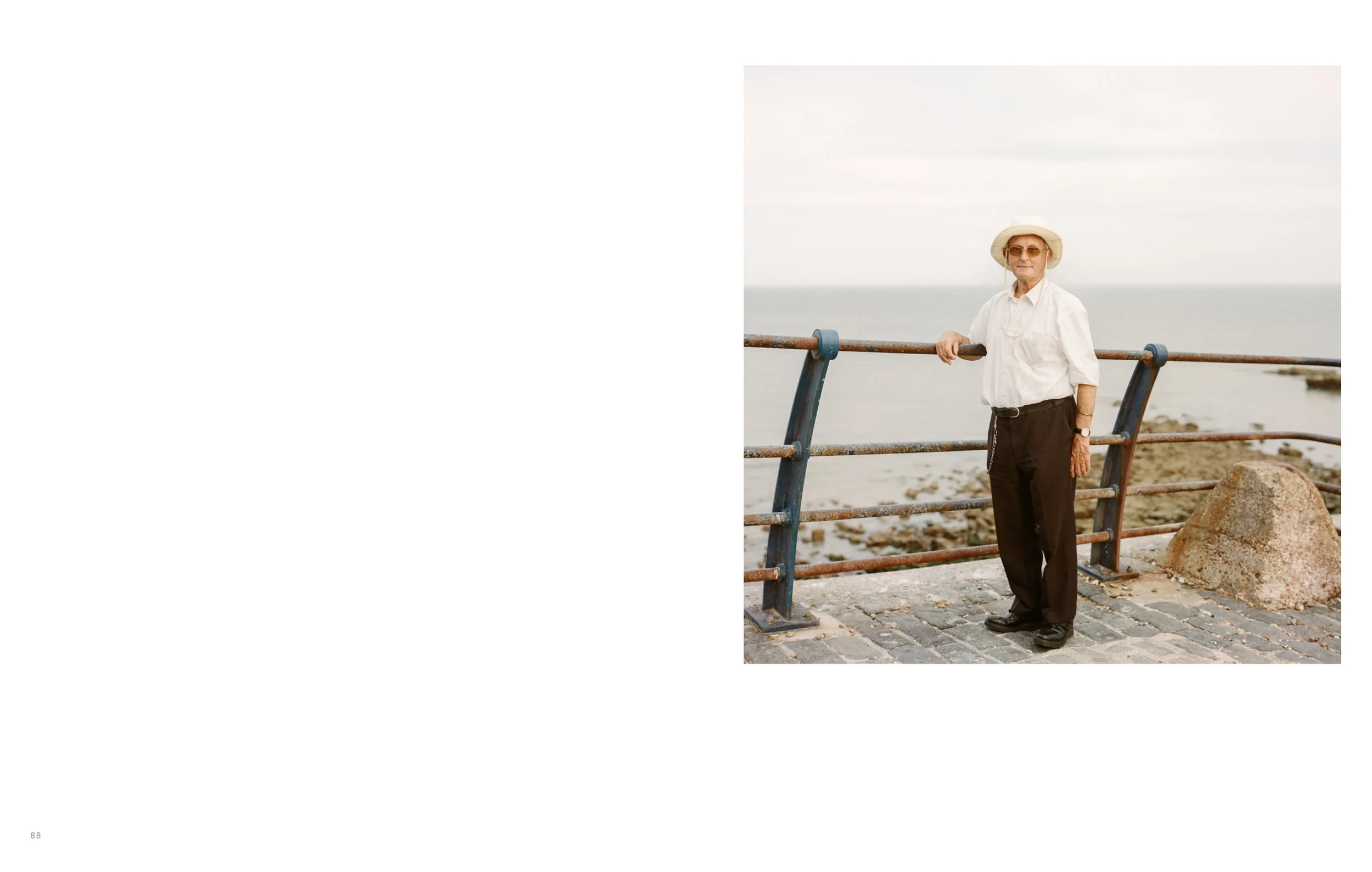 An elderly man wearing a white shirt, black pants, and a wide-brimmed hat standing on a pier, holding onto a rusty metal railing with the ocean in the background.