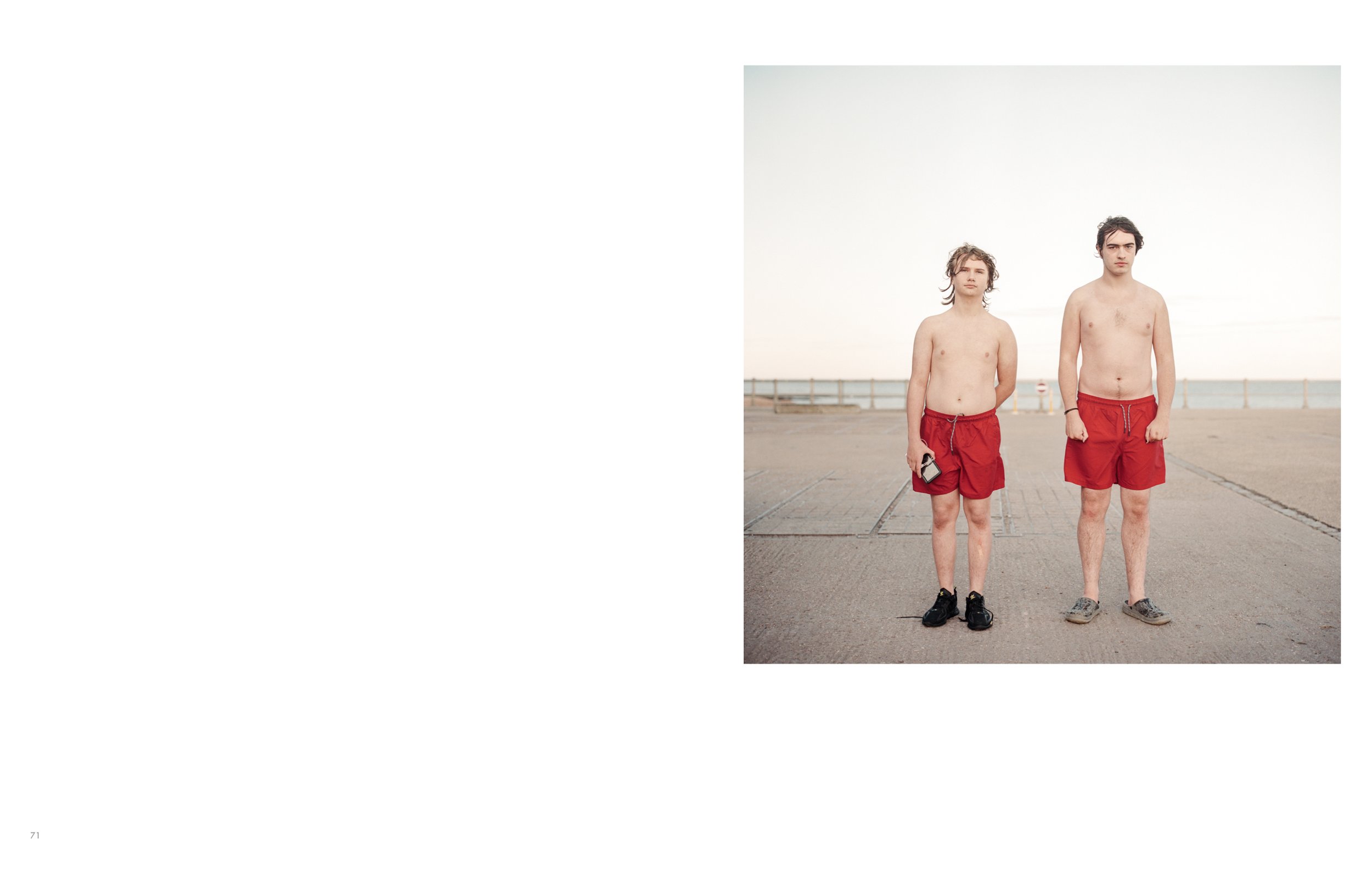 Two young men stand shirtless on a paved area near the coast, wearing red shorts and black sneakers, with a wooden fence and the ocean in the background.