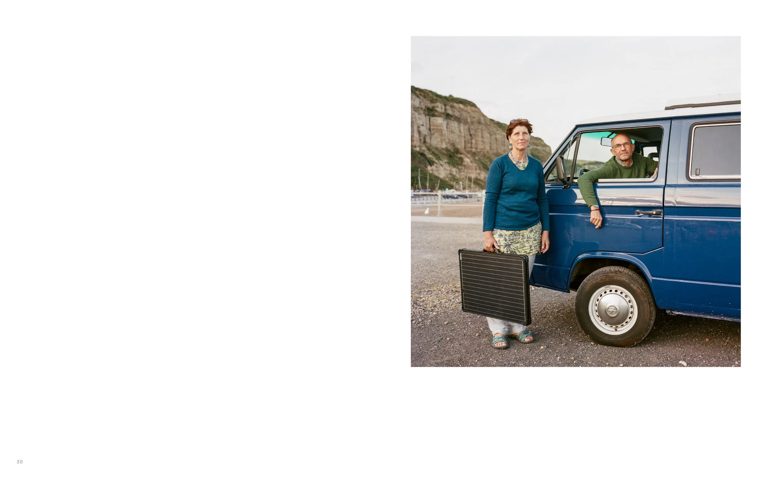 An older woman standing next to a blue van, holding a black briefcase, and a man inside the van leaning out of the window, parked near a beach with cliffs in the background.