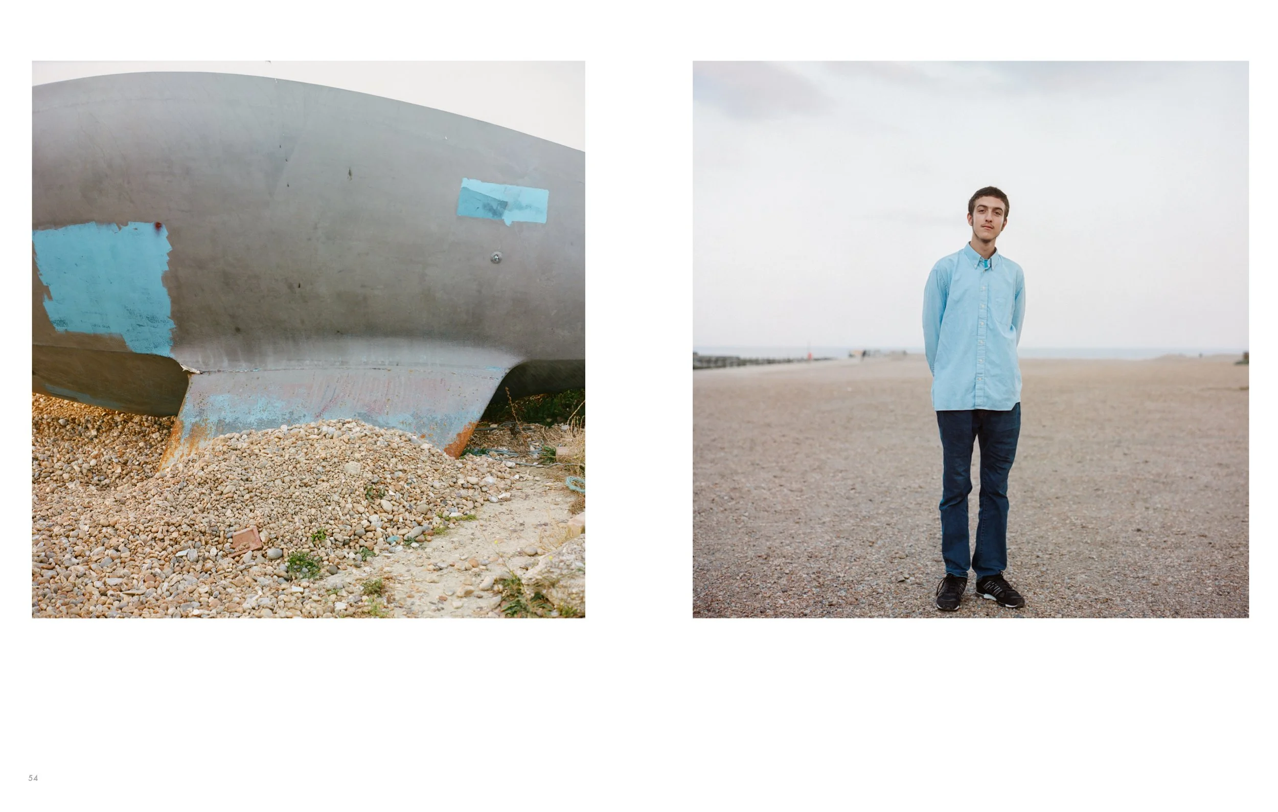 A boat on gravel next to a person standing on the beach. The boat is weathered with patches of blue paint. The person is wearing a light blue shirt, dark pants, and sneakers, standing with hands behind their back against a cloudy sky.