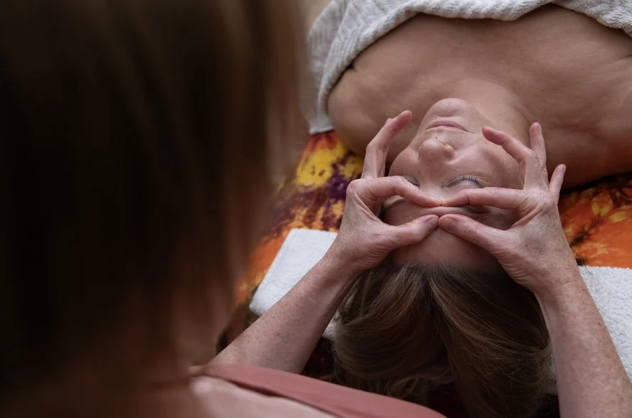 A person giving a facial massage to a woman lying on her back, outdoors on a colorful blanket.