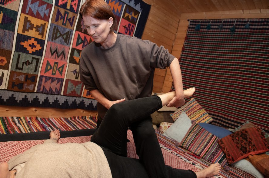 A woman receiving a massage or physical therapy from another woman in a room decorated with colorful, patterned textiles on the walls and cushions on the floor.