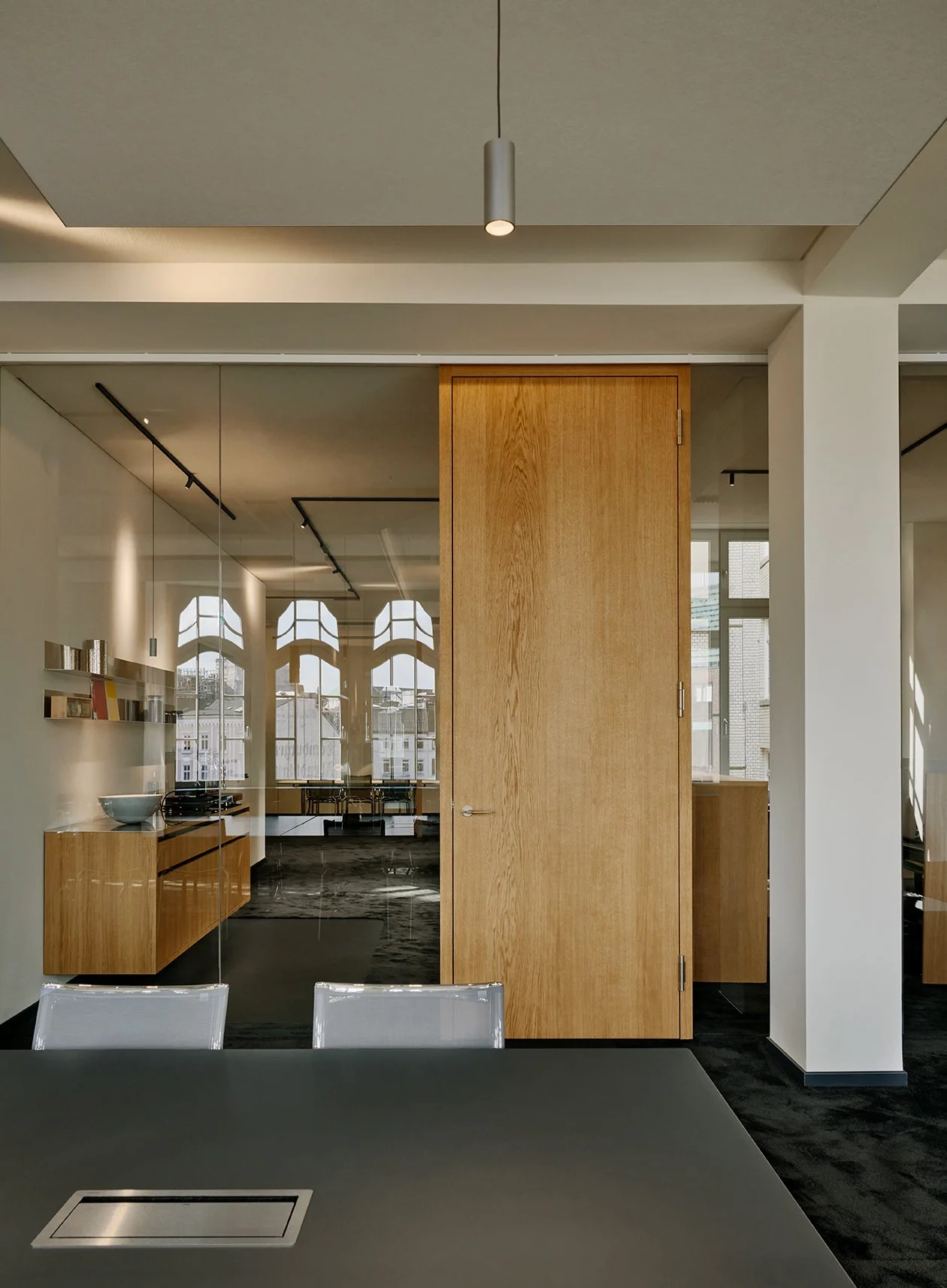 Innenraum eines modernen Büros mit Holz- und Glaselementen, einem Tisch und Stühlen, sowie großen Fenstern für natürliches Licht.