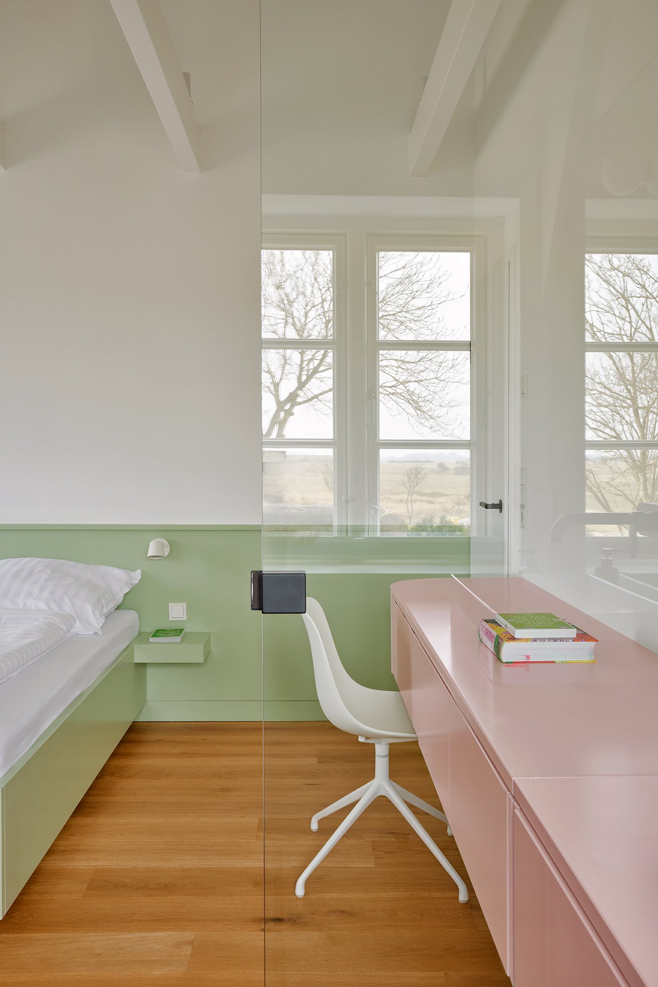 Blick in ein modernes, helles Schlafzimmer mit einer großen Fensterfront, einem Doppelbett, einer Nachttischlampe, einem Schreibtisch mit einem weißen Drehstuhl und einem Bücherstapel auf dem Tisch.