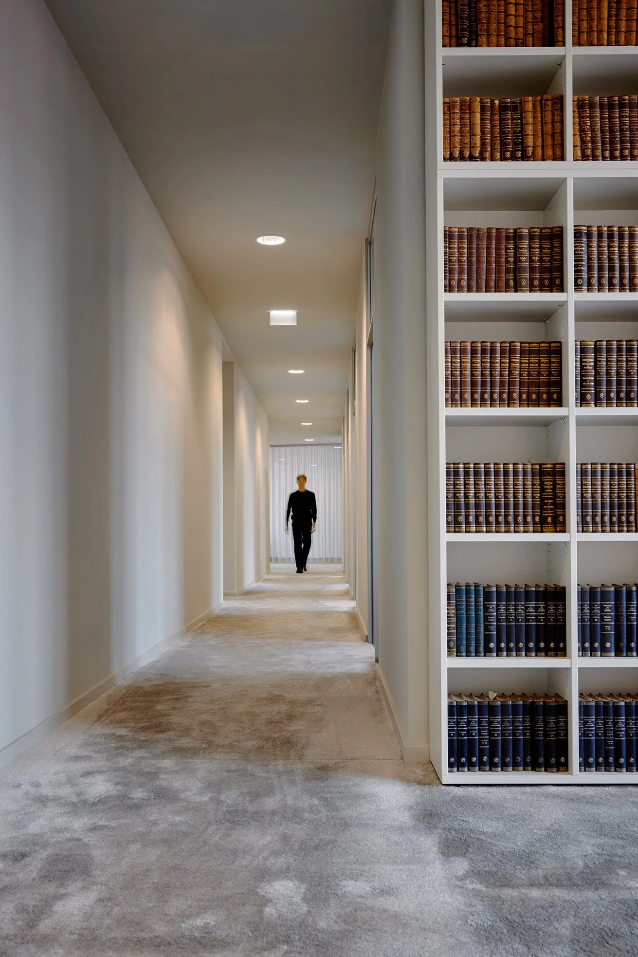 Bücherschrank in einem Bibliotheksflur, im Hintergrund eine Person auf dem Flur, der mit Deckenleuchten beleuchtet ist.