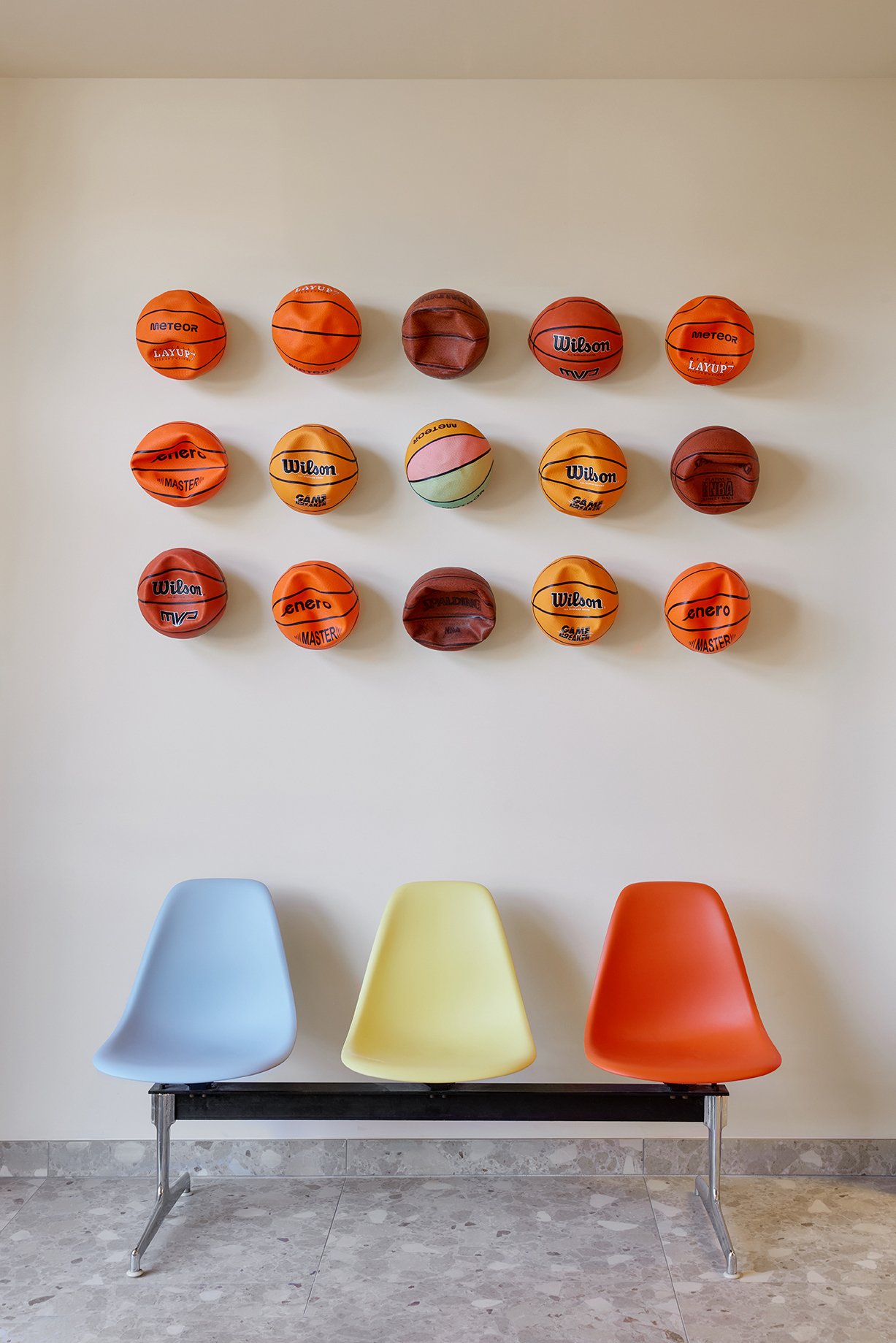 Eine Wand mit 15 Basketballbällen in verschiedenen Farben und Marken, darunter Wilson und Meteor. Darunter befinden sich drei Stühle in den Farben Blau, Gelb und Rot. Der Boden ist hellgrau mit weißen und schwarzen Mustern.