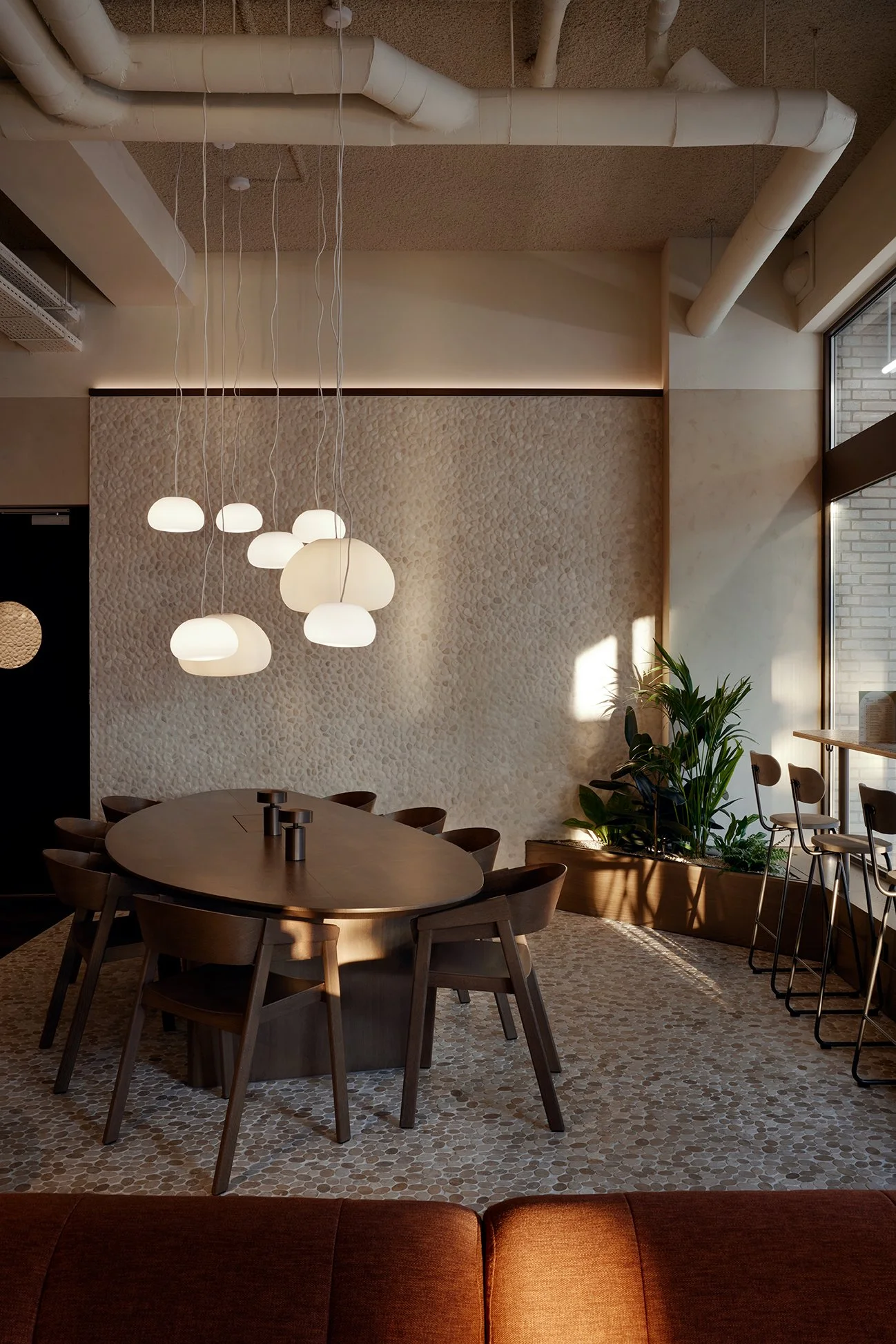 Innenansicht eines modern eingerichteten Cafés oder Restaurants mit einem runden Holztisch, Stühlen, Pendelleuchten über dem Tisch, einer Pflanzenpflanzung, großen Fenstern und einer Wand mit Mosaikfliesen.