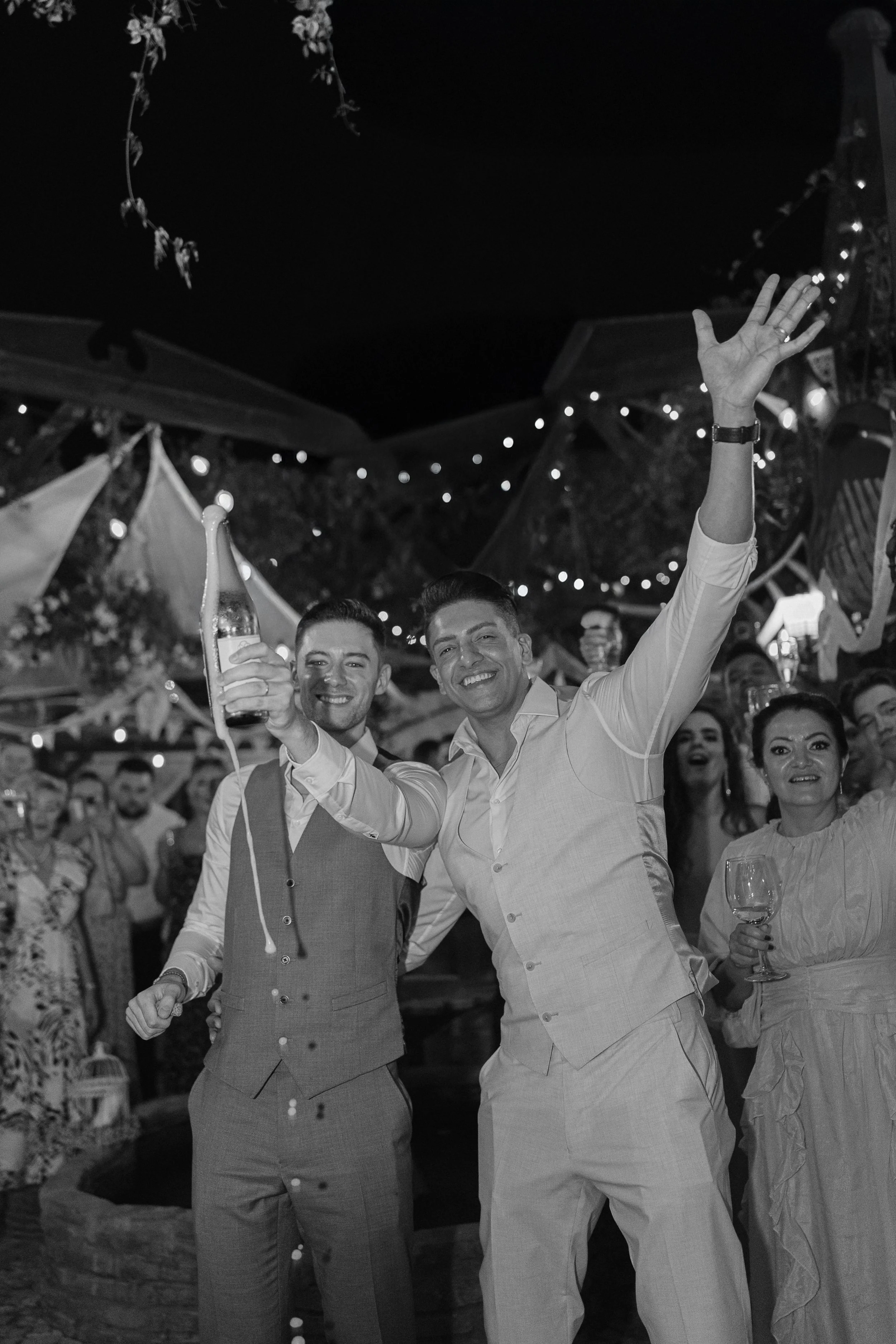 A bride and groom walking together outdoors on their wedding day, with the bride holding a bouquet of flowers and touching the groom's face. There is a vintage car behind them and lush greenery around.