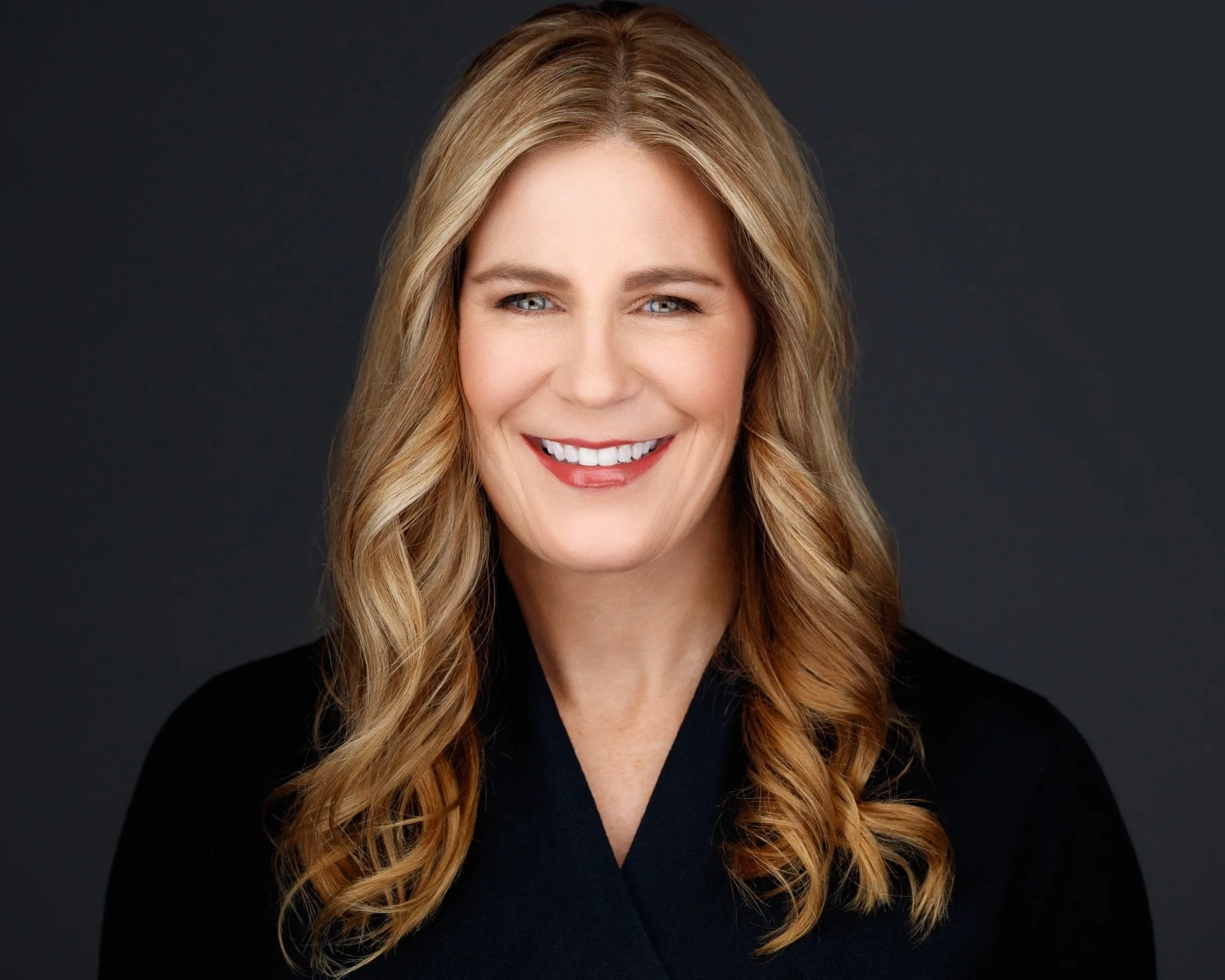 A smiling woman with wavy blonde hair wearing a black top against a dark background.