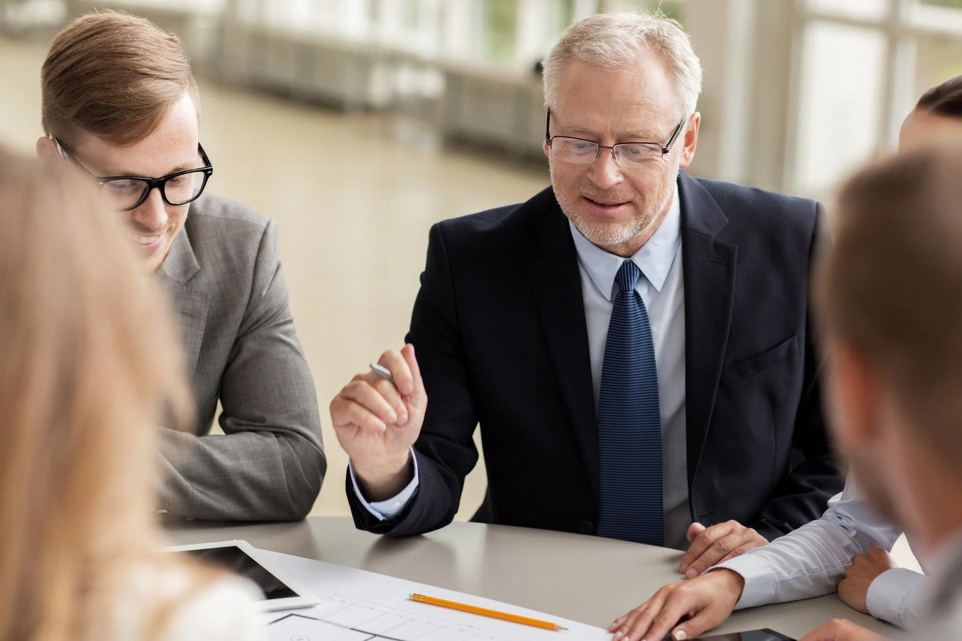 A group of young professionals and an older man engaged in a meeting around a table, with papers and a tablet in front of them.
