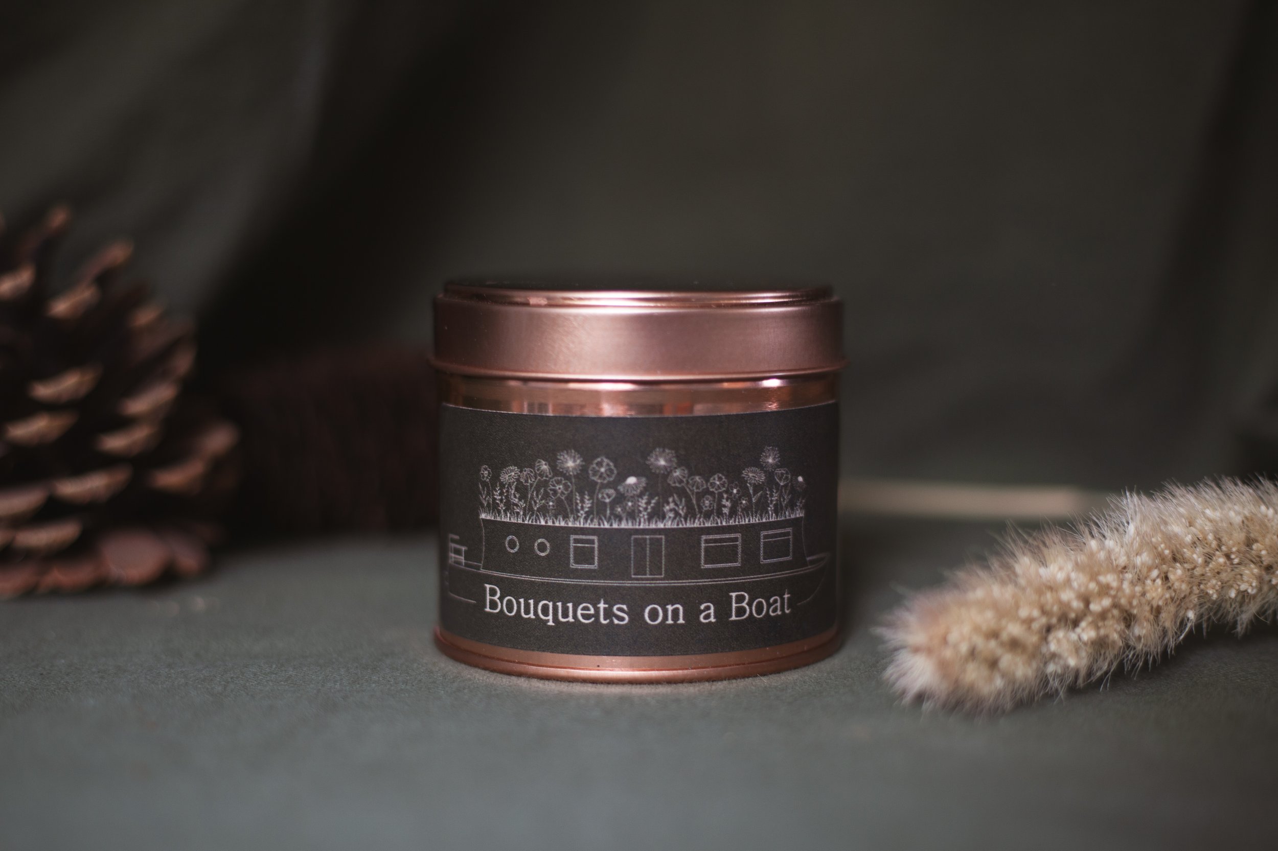 Bouquets on a Boat organic aromatherapy soy candle in an amber glass jar with a copper lid, styled with dried grasses and pinecones on a soft green fabric background.