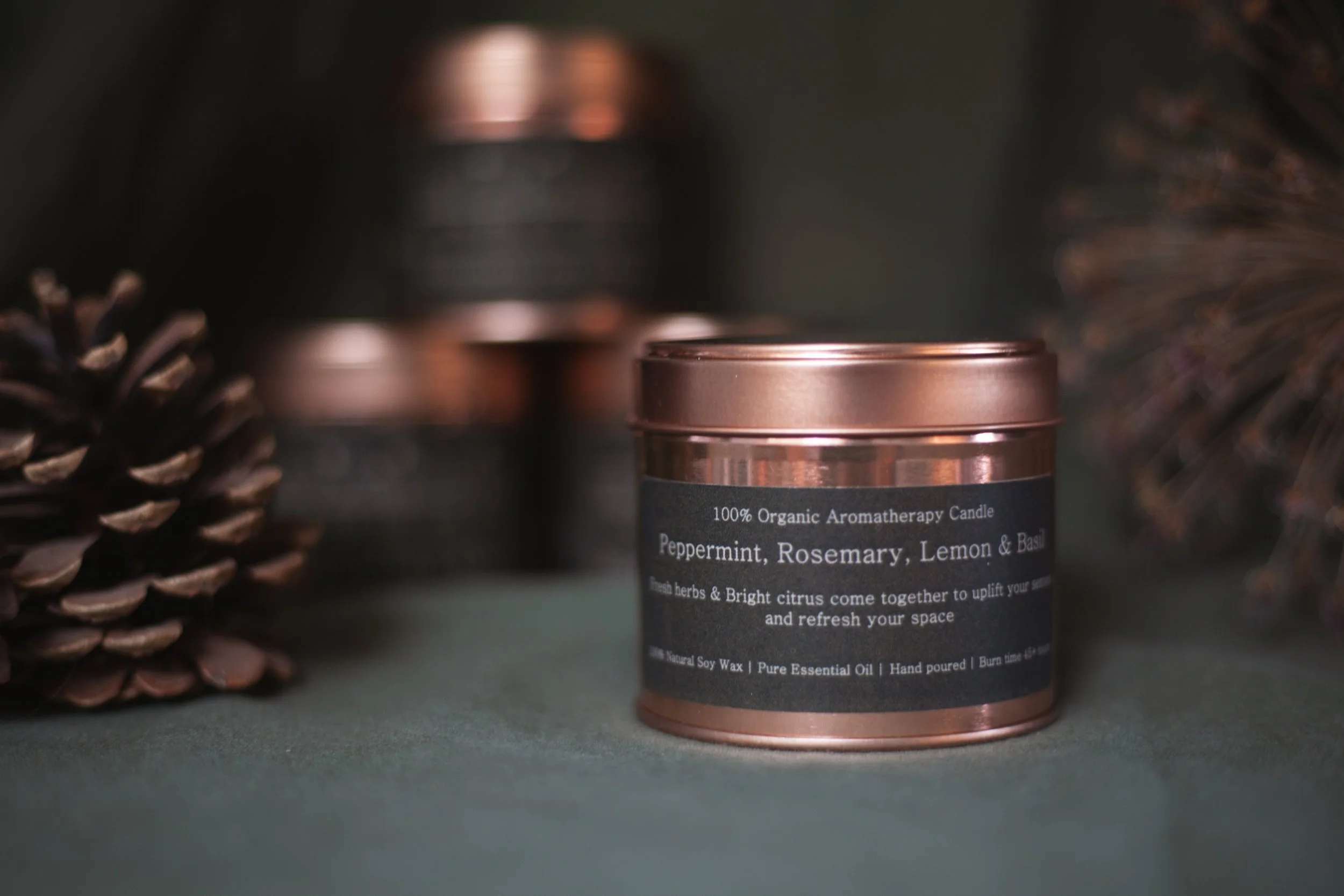 Rosemary, Peppermint, Lemon and Basil organic aromatherapy soy candle by Bouquets on a Boat in an amber glass jar with a copper lid, styled with pinecones and dried flowers.