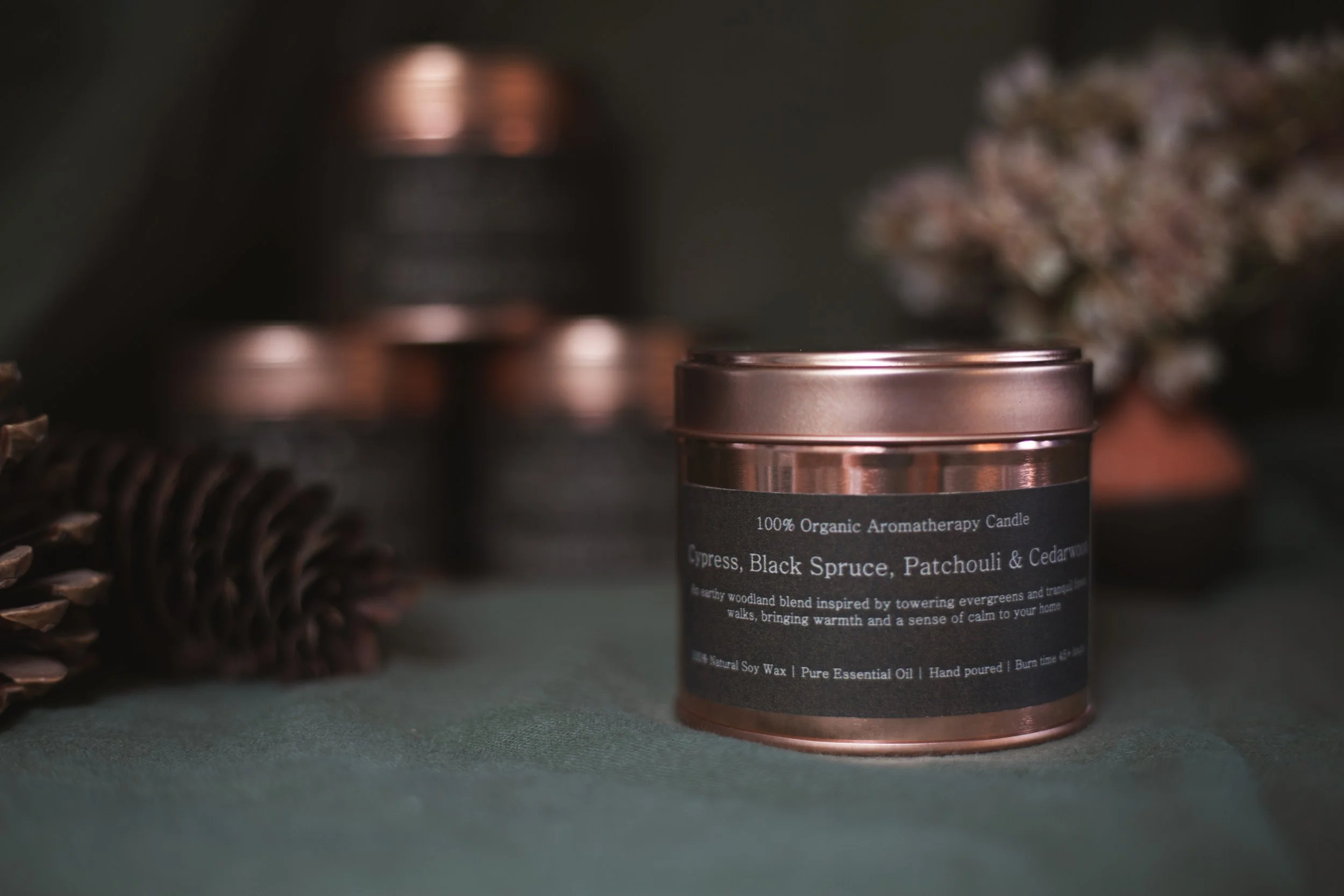 Cypress, Black Spruce, Patchouli & Cedarwood organic aromatherapy soy candle by Bouquets on a Boat in an amber glass jar with a copper lid, styled with pinecones and dried flowers.