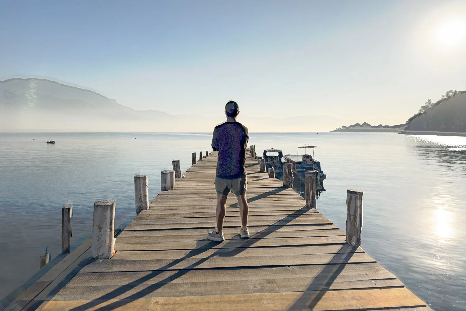 Abstract ICM Bluristic  photo - Person standing on a wooden pier looking out over a calm body of water with distant mountains, boats docked nearby, and the sun shining in a clear sky.