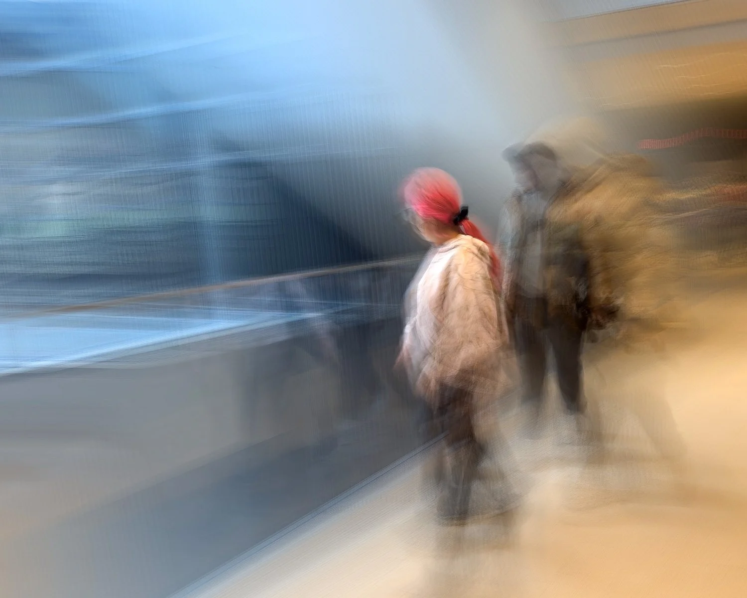 Abstract ICM Bluristic  photo - Blurred image of two people walking on a street, one person wearing a pink headscarf and beige coat, the other in darker clothing, with a cityscape in the background.