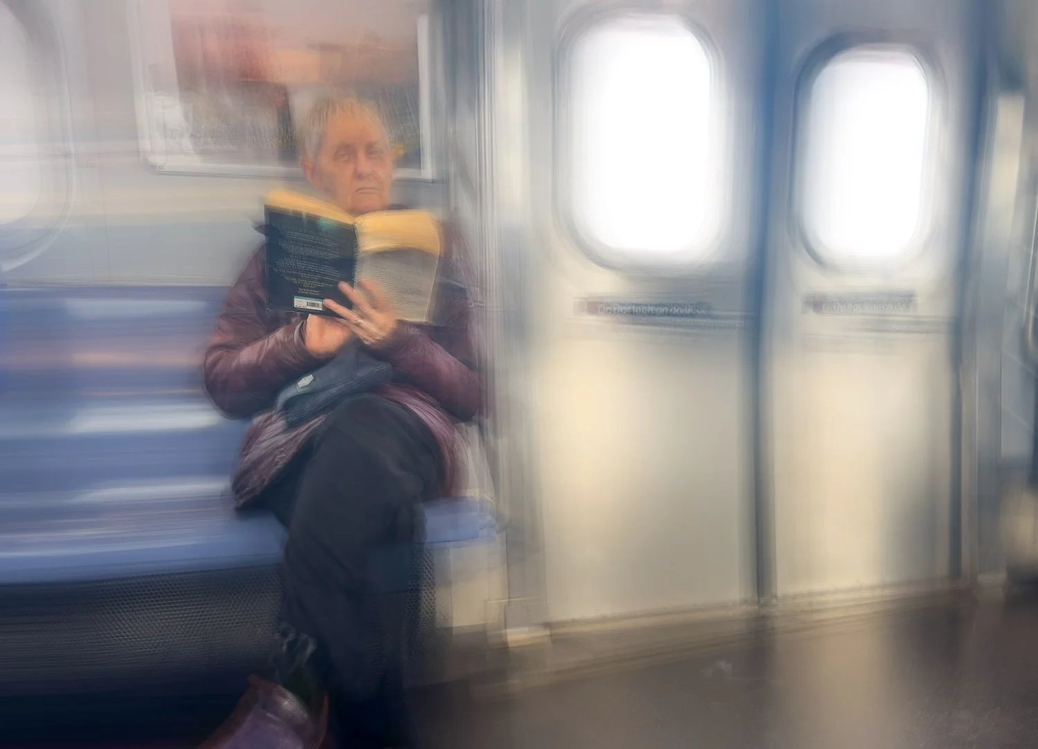Abstract ICM Bluristic  photo - A person sitting on a subway train reading a book, with motion blur from the train's movement.