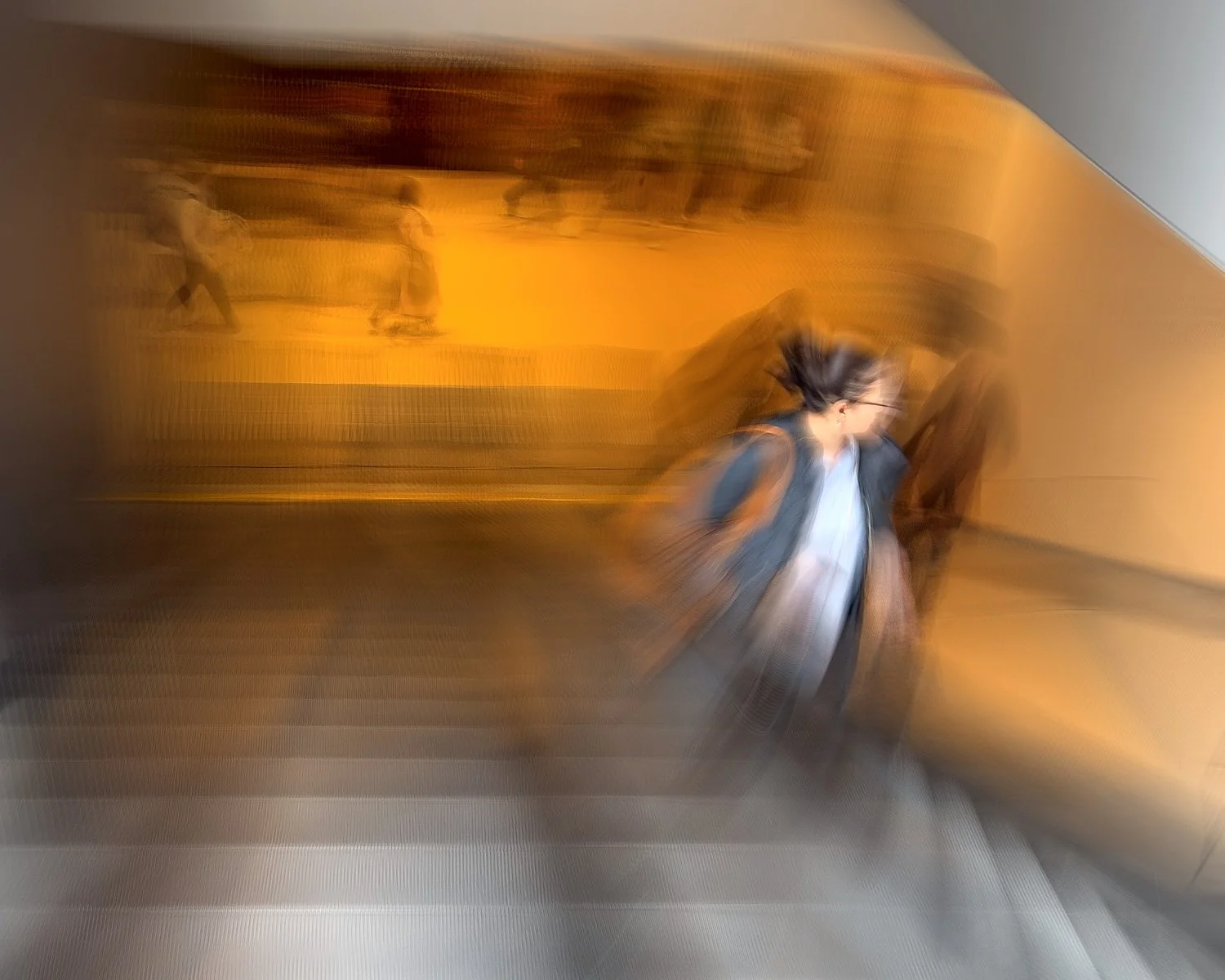 Abstract ICM Bluristic  photo - A person with glasses and a backpack walking down an escalator, with an orange-lit background and other people in the distance, all appearing blurred due to motion.