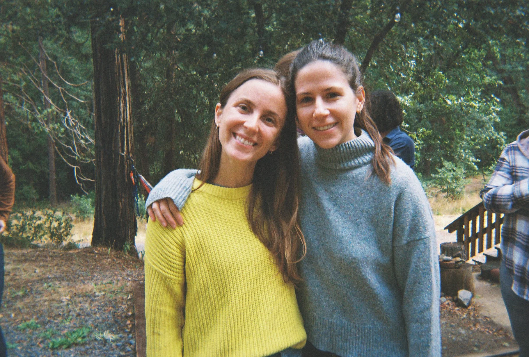 Two women with long hair smiling and standing closely together outdoors in a park, with one wearing a yellow sweater and the other in a gray sweater, in front of trees, a wooden fence, and other people in the background.