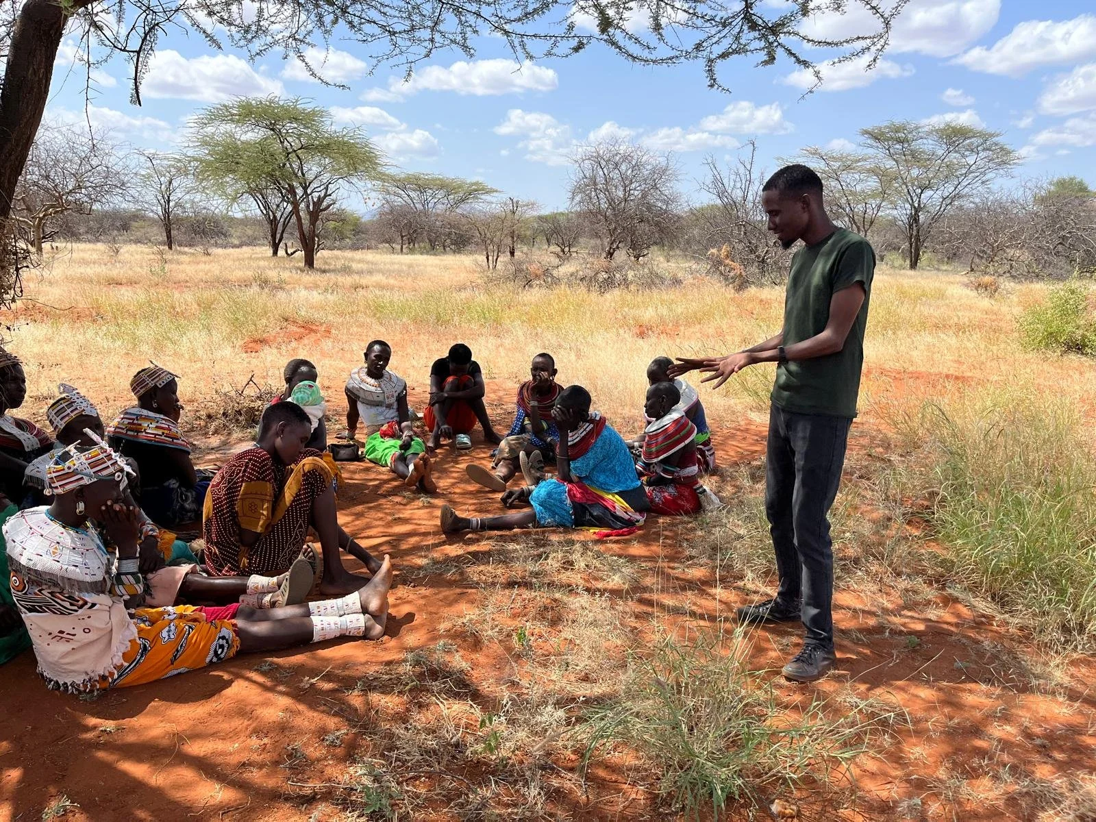 A group of people in Kenya sitting on the ground in a semi-circle outdoors under a tree, with a man standing and speaking to them in a dry, open landscape.