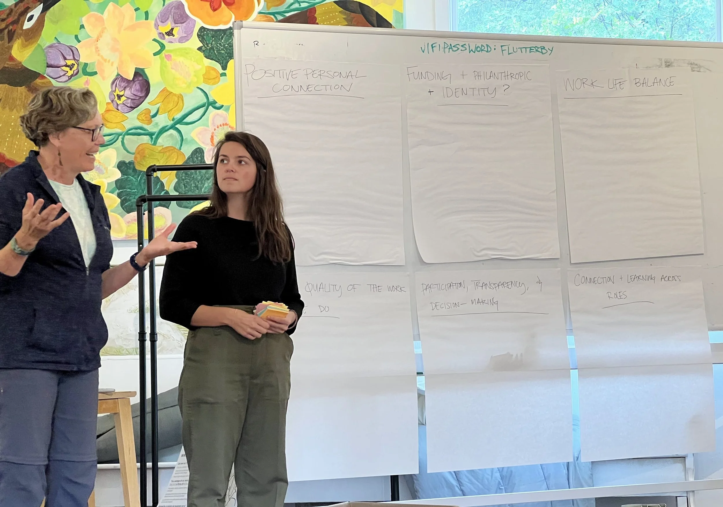 Two women standing in front of whiteboards filled with handwritten notes, with a colorful floral mural on the wall behind them.