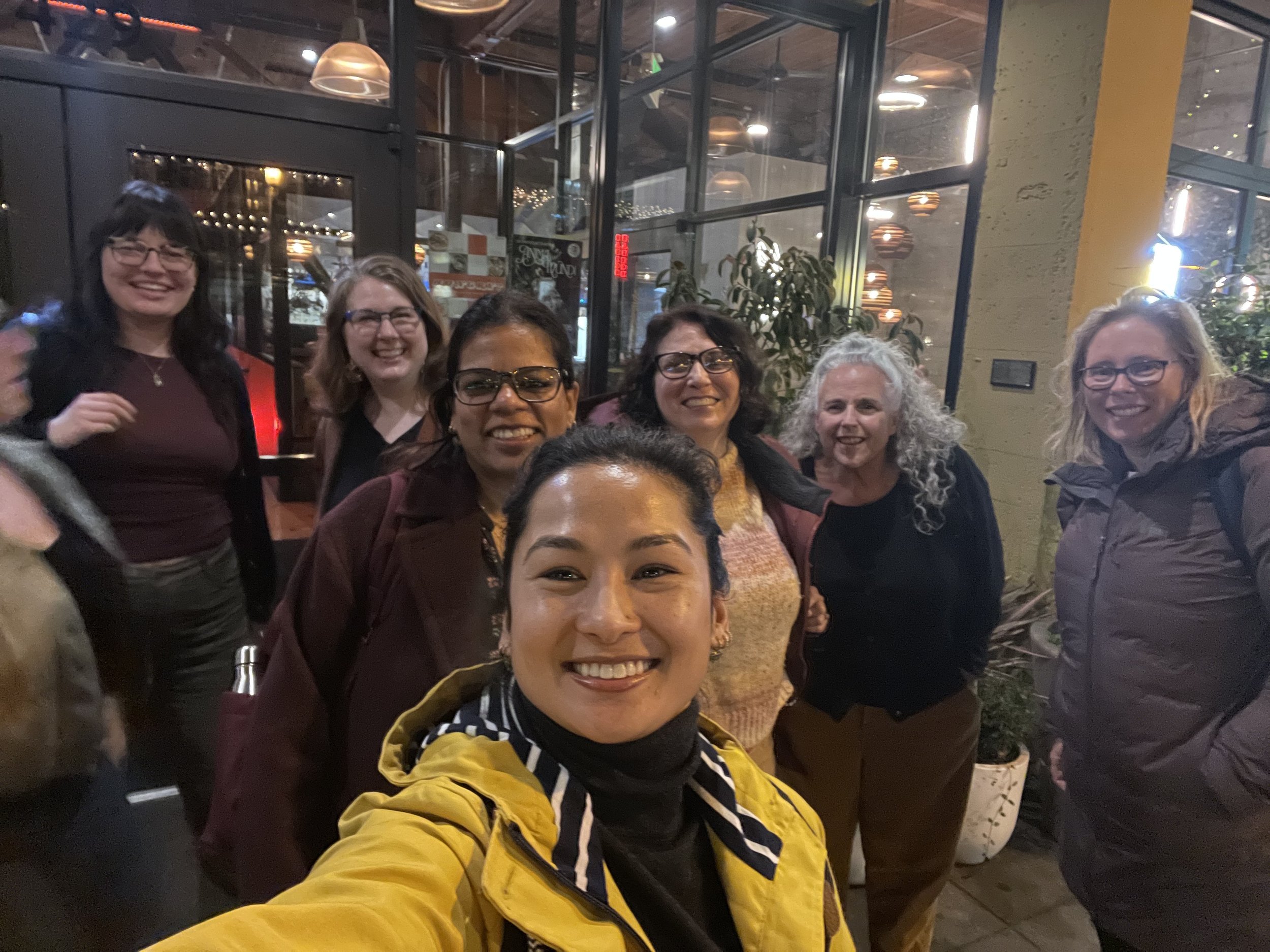 A group of six women smiling and posing for a selfie inside a restaurant with large glass windows and hanging lights.