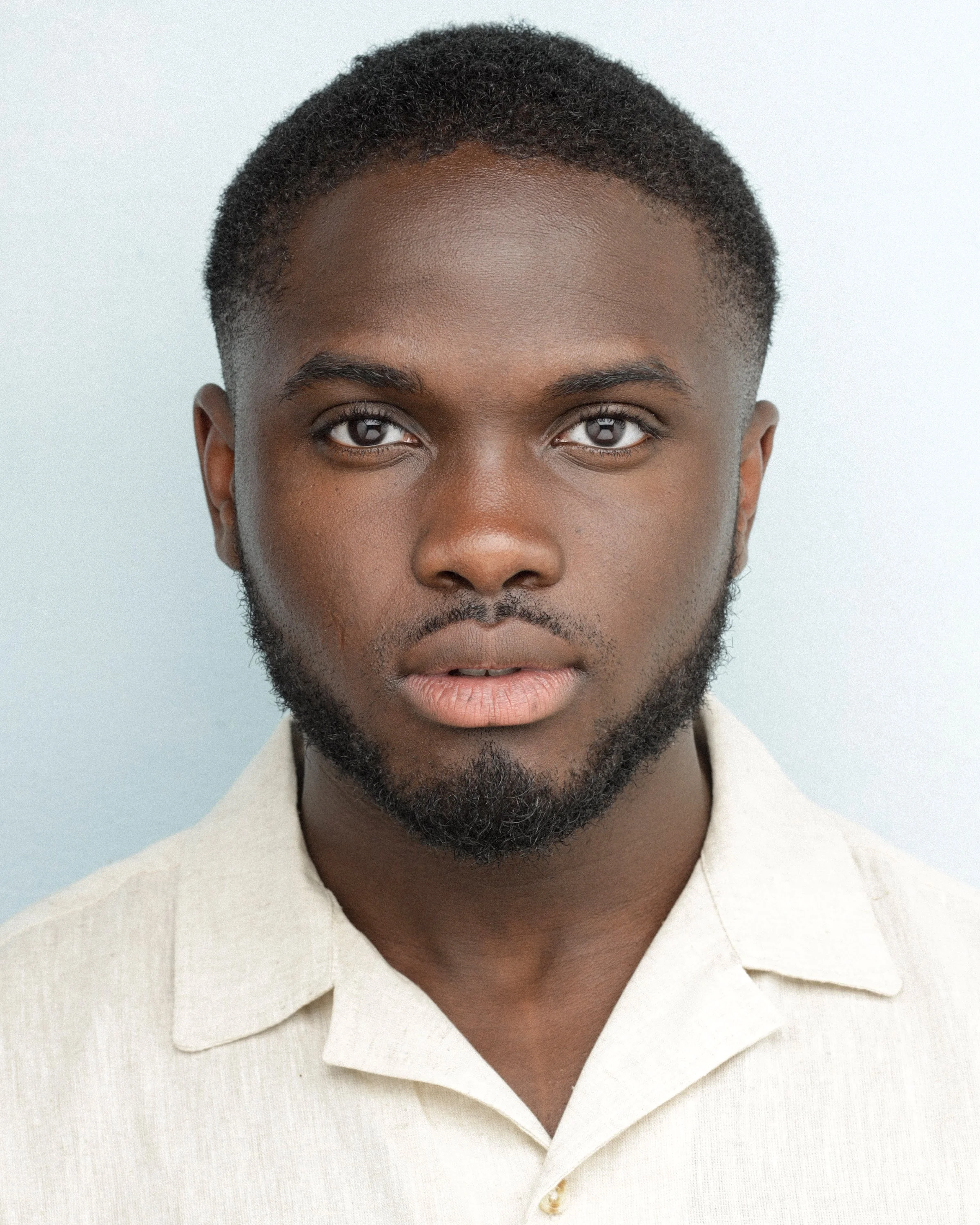 Professional headshot of London Portrait Photographer Donnell Asare, featuring short curly hair, a neatly groomed beard, and a light collared shirt against a plain background, captured in soft, even lighting to highlight facial features.