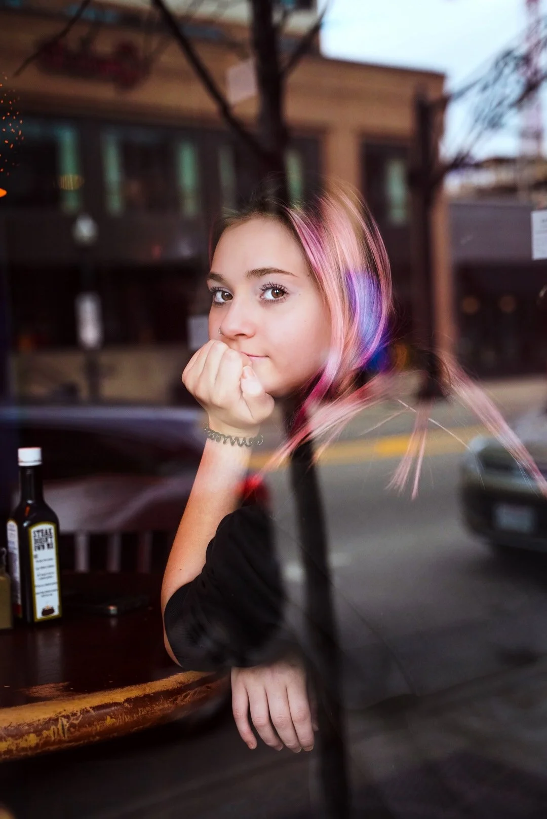 Girl with pink hair sitting at a table by a window, looking outside, with a bottle on the table.