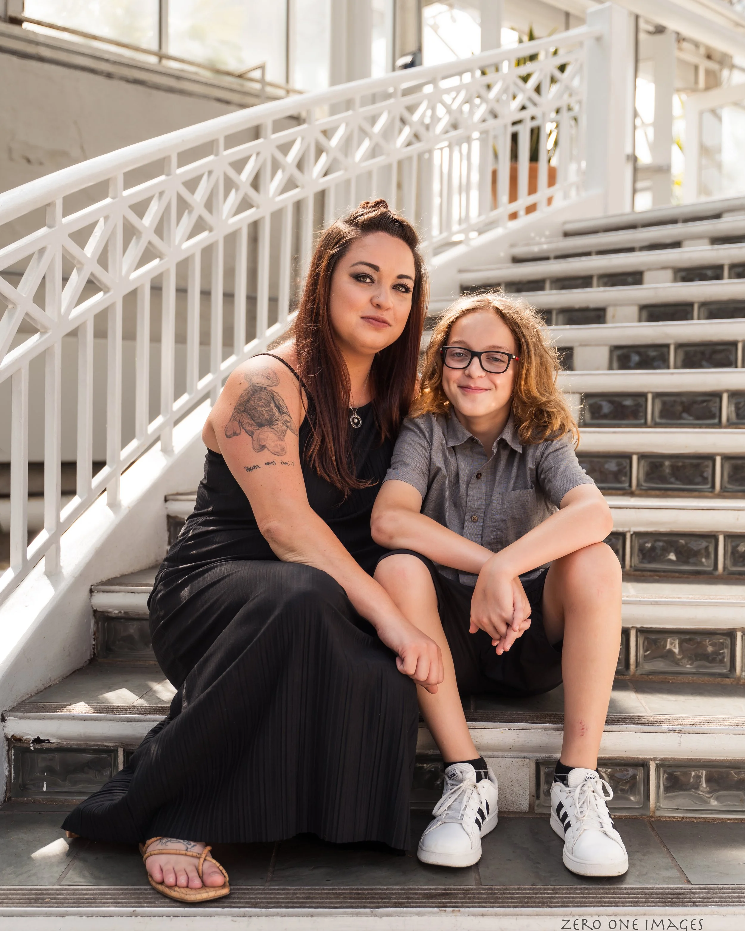 A woman and a boy sitting on stairs indoors, with a white railing behind them. The woman has long brown hair, a tattoo on her arm, and is wearing a black dress and sandals. The boy has long hair, glasses, and is wearing a gray shirt, black shorts, and white sneakers.