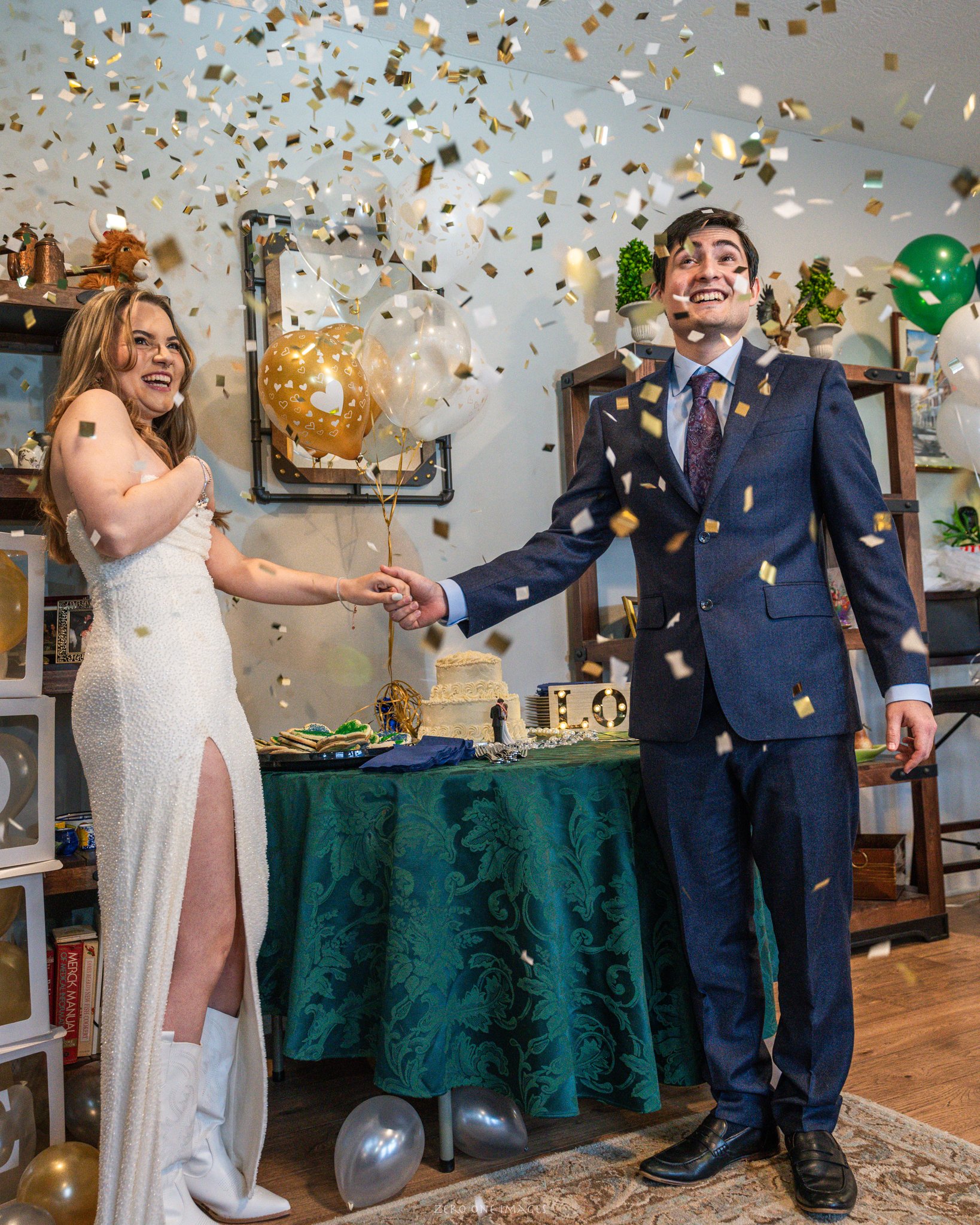 A woman in a white dress and a man in a suit celebrating indoors with balloons, confetti, and cake on a decorated table.