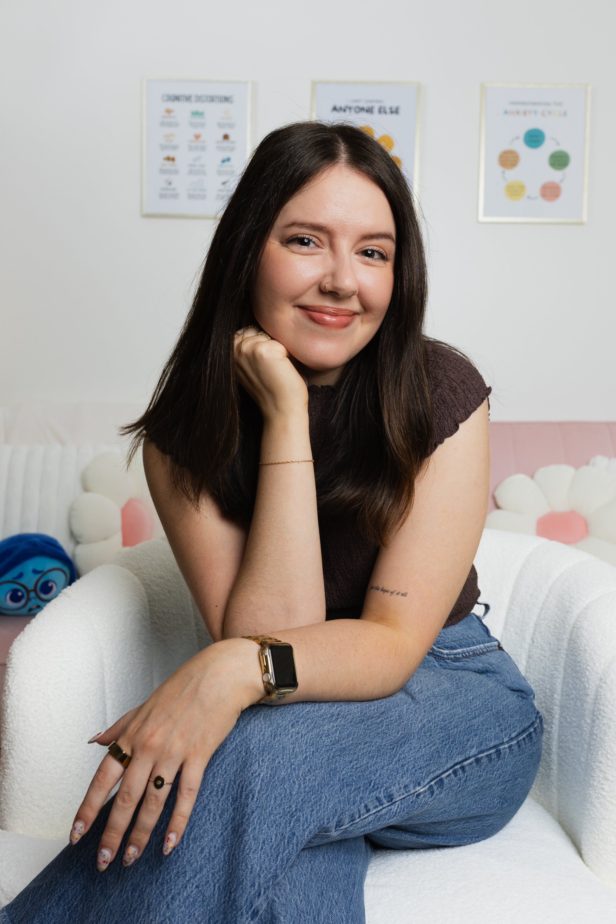 A young woman with long dark hair, wearing a dark top and jeans, sitting on a white plush chair in a room with minimalist decor and framed posters on the wall behind her. She has a gold ring, a watch, and a tattoo on her forearm, and she is smiling with her hand resting on her chin.