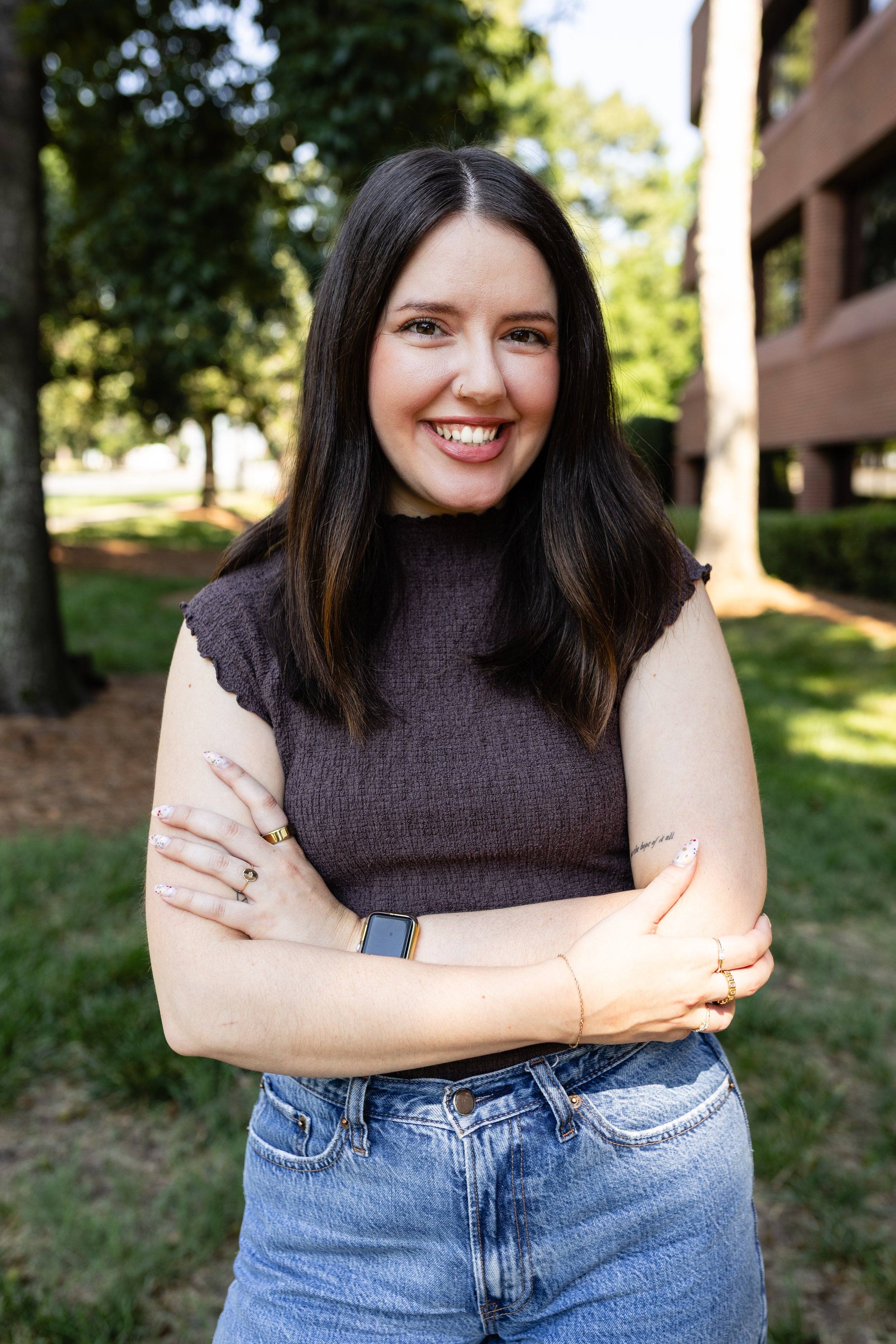 A young woman with long dark hair smiling outdoors on a sunny day, crossing her arms, wearing a dark sleeveless top, blue jeans, and a wristwatch, with trees and a building in the background.
