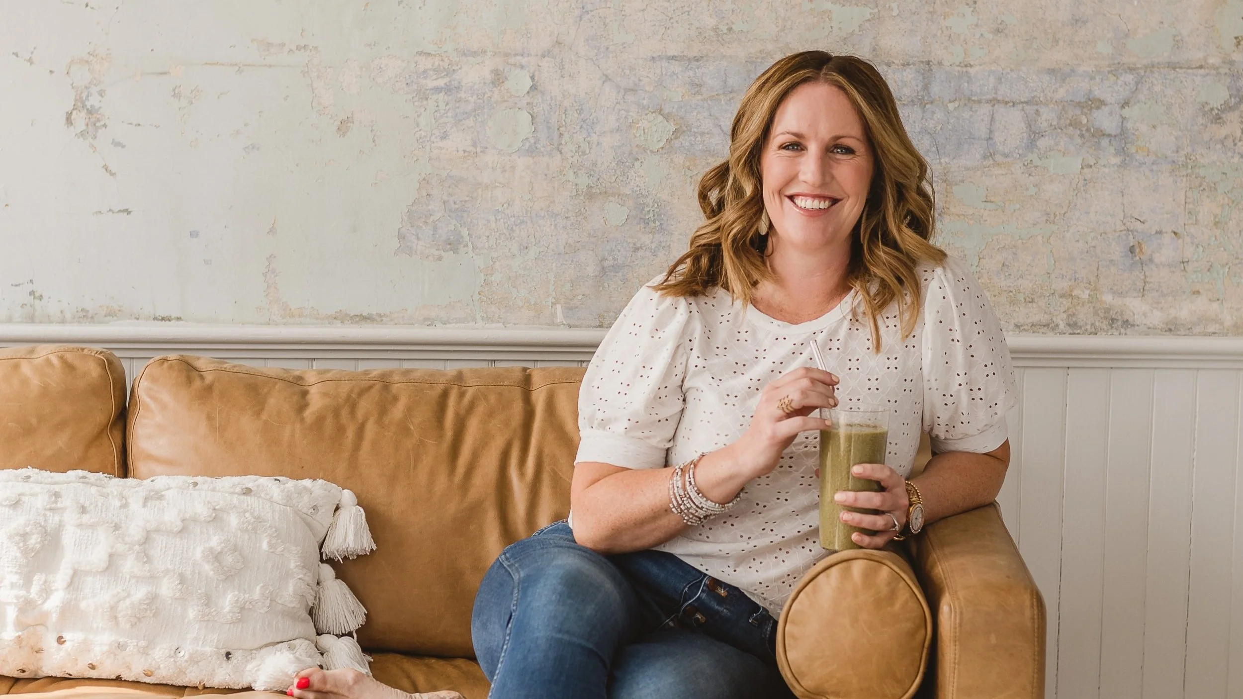 A woman sitting on a tan leather couch, holding a cup of green smoothie, smiling at the camera, in a cozy room with textured walls and white paneling.