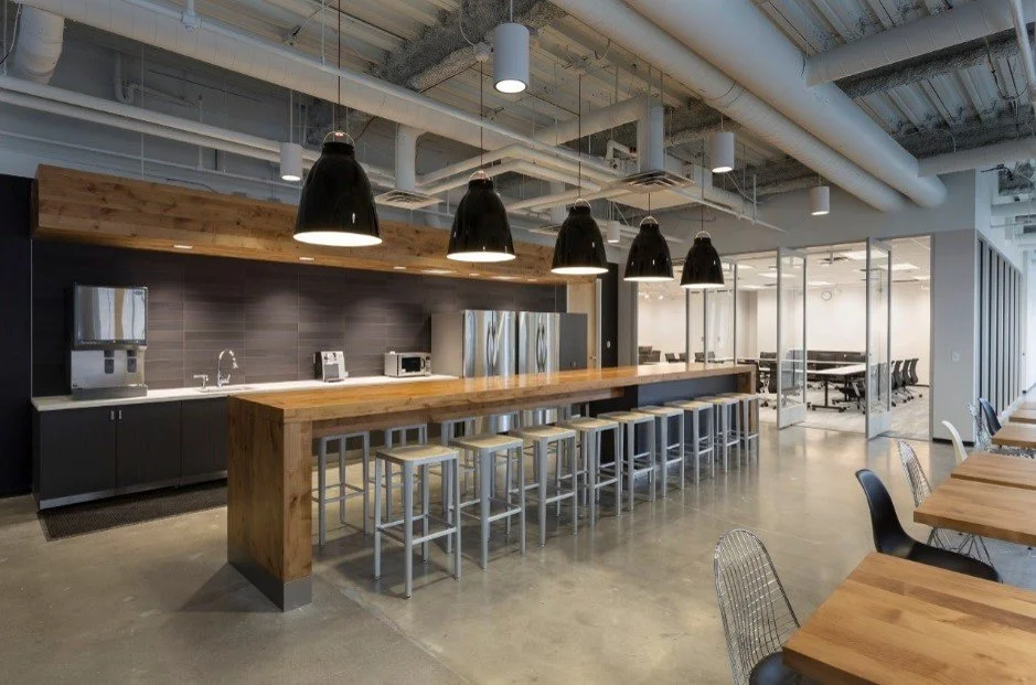 Modern office break room with a long wooden counter, black pendant lights, and high metal stools. Kitchen area with microwave, water dispenser, and refrigerator. Adjacent glass-walled conference room with black chairs and tables.