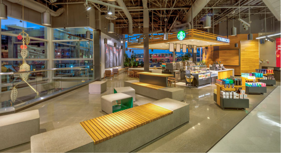 Interior of a modern airport with a Starbucks coffee stand, seating area, and gift shop with colorful items.