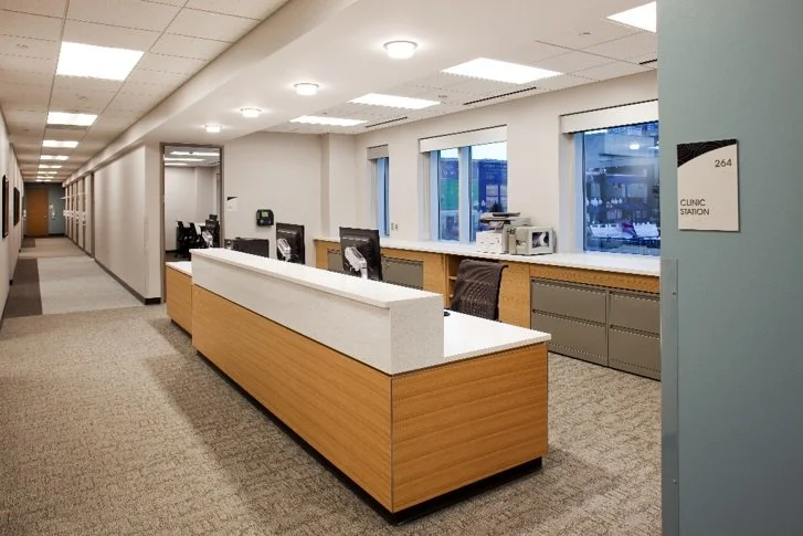 Empty clinic reception area with a long wooden counter, computers, chairs, and large windows in a medical facility.