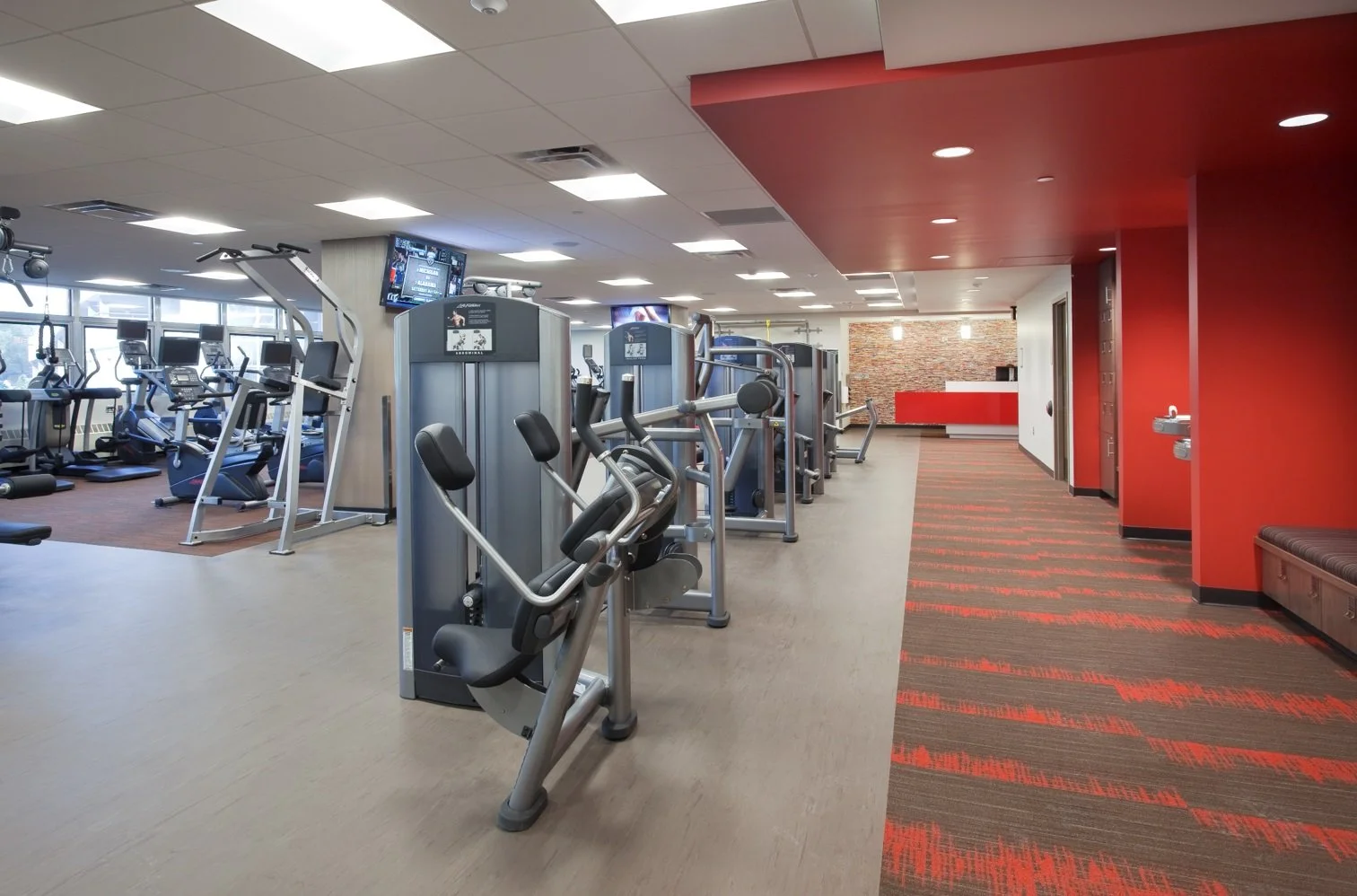 Empty gym with various workout equipment including treadmills, elliptical machines, and weight machines, with a red accent wall and a reception area in the background.