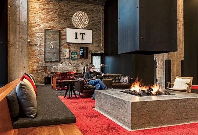 Interior of a modern lounge with a brick wall, eclectic furniture including a leather sofa, a fireplace, and a person sitting on the sofa using a laptop. Decor includes framed artwork, a chandelier, and a large sign that reads 'IT'.