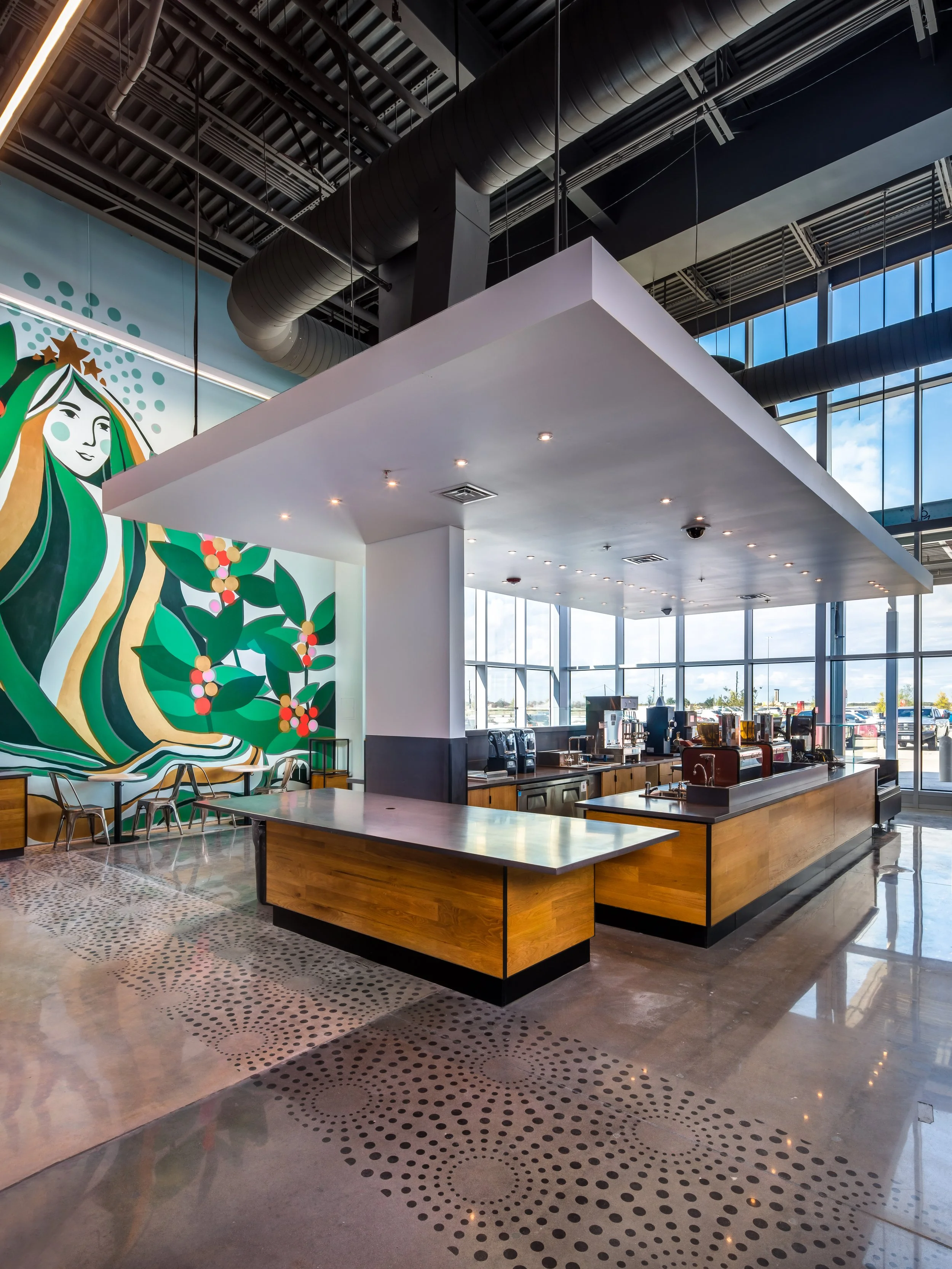 Modern coffee shop interior with wood counters, coffee machines, large windows, and colorful mural on the wall.