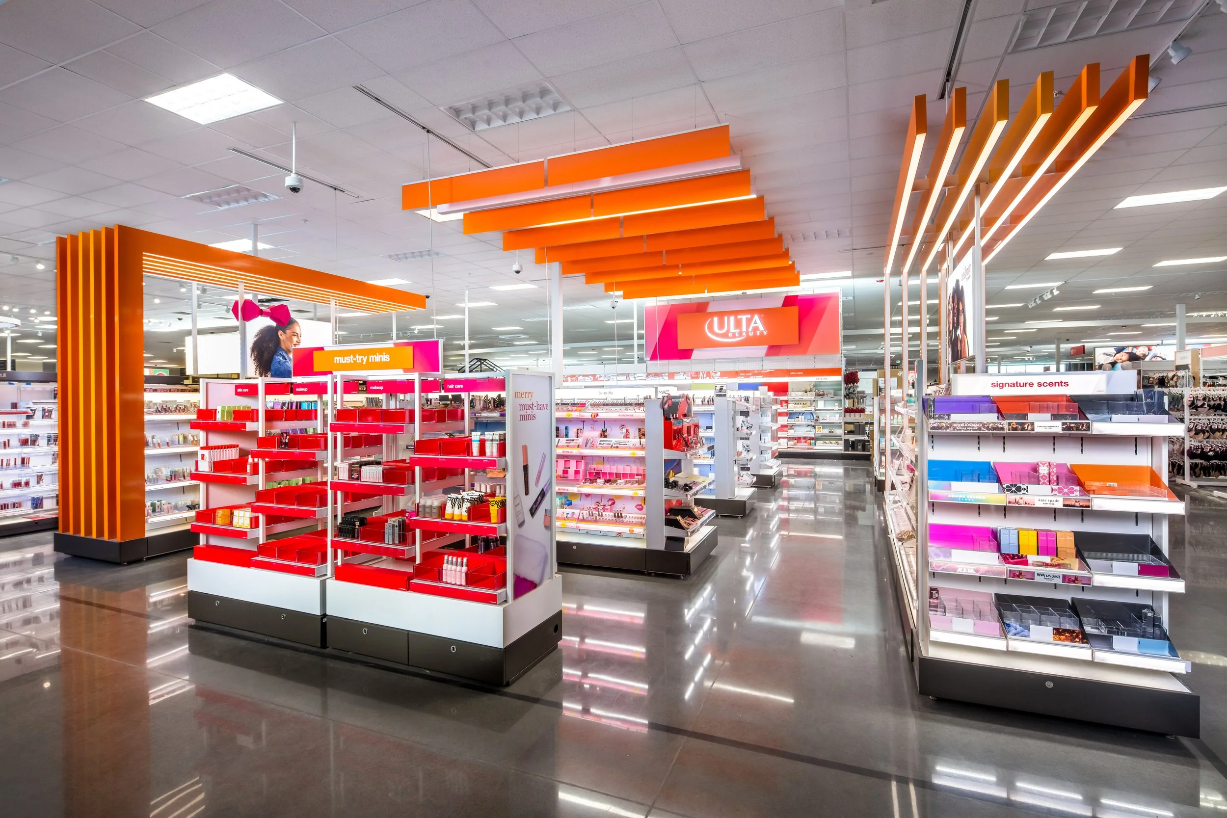 Inside a retail store with bright lighting, displays of makeup, cosmetics, and gift sets, featuring orange and pink accents and a large ULTA sign in the background.